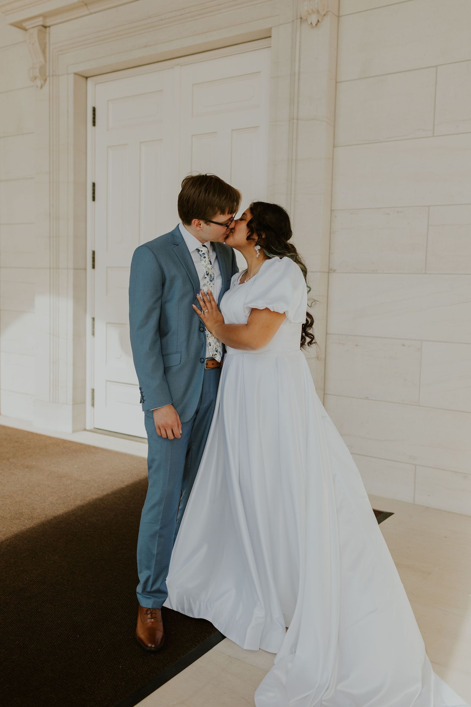 A couple in formal wedding attire shares a tender moment against a white paneled wall.