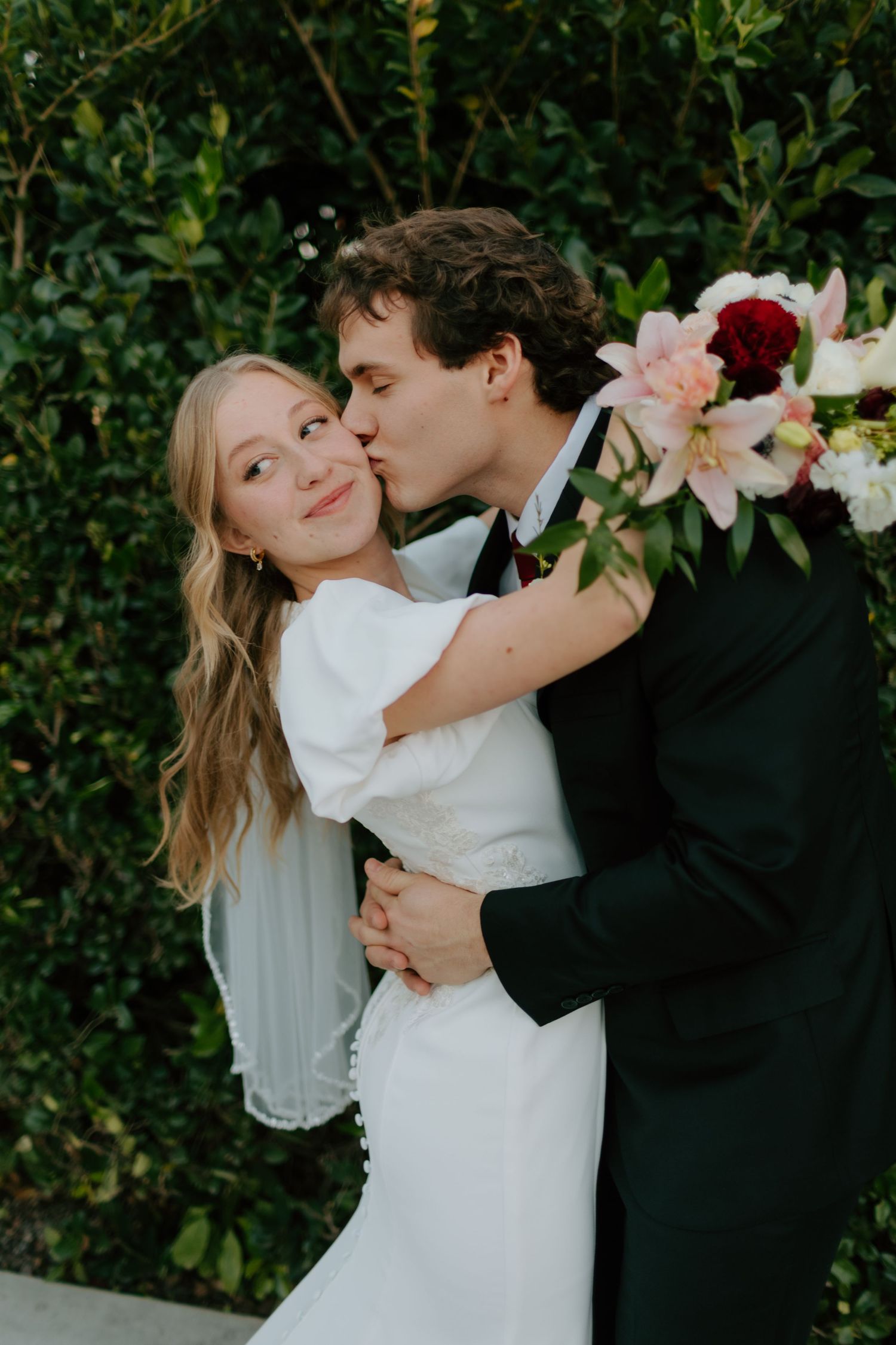 Groom in black tuxedo kissing bride on cheek while embracing her holding pink and red bouquet.