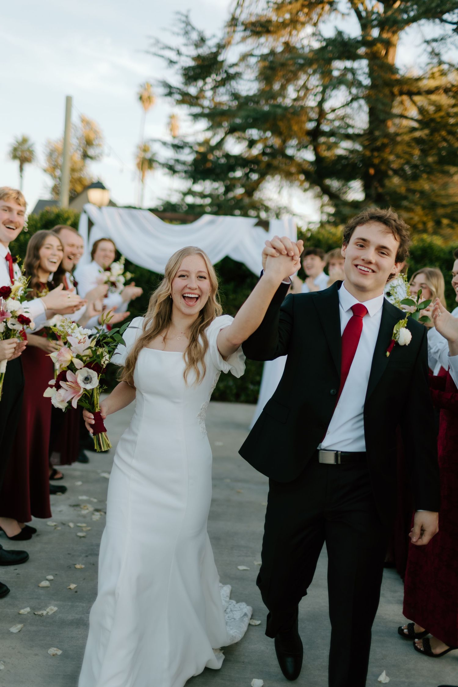 Joyful newlyweds raise hands walking past wedding party in burgundy, bride in white gown, groom in black suit with red tie.