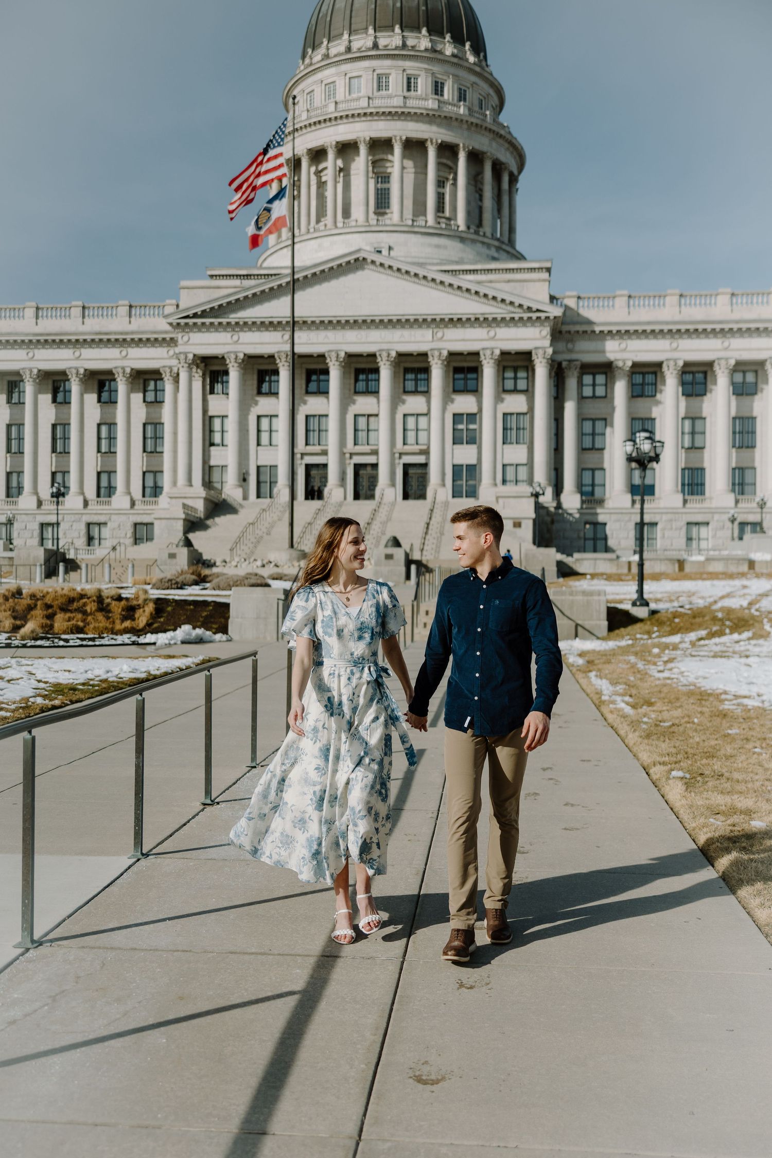 Couple holding hands and walking in front of the Utah State Capitol building on a sunny winter day.