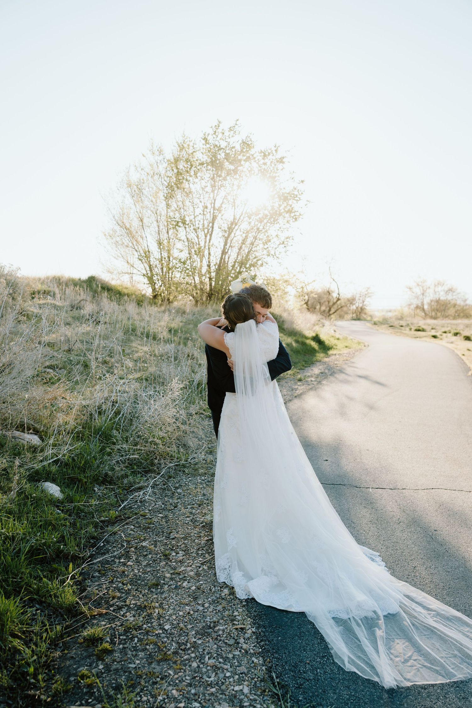 Bride and groom embrace on a sunlit road surrounded by nature in romantic wedding photo.