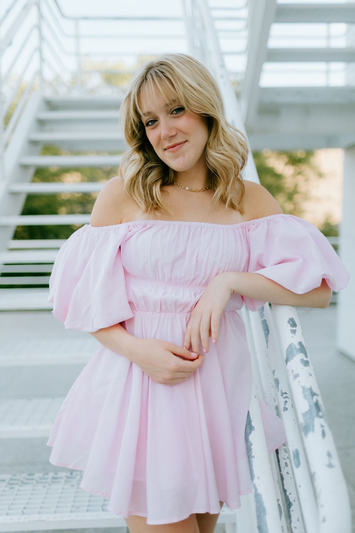 Young blonde woman in pink puff-sleeve off-shoulder mini dress posing near white stairs.