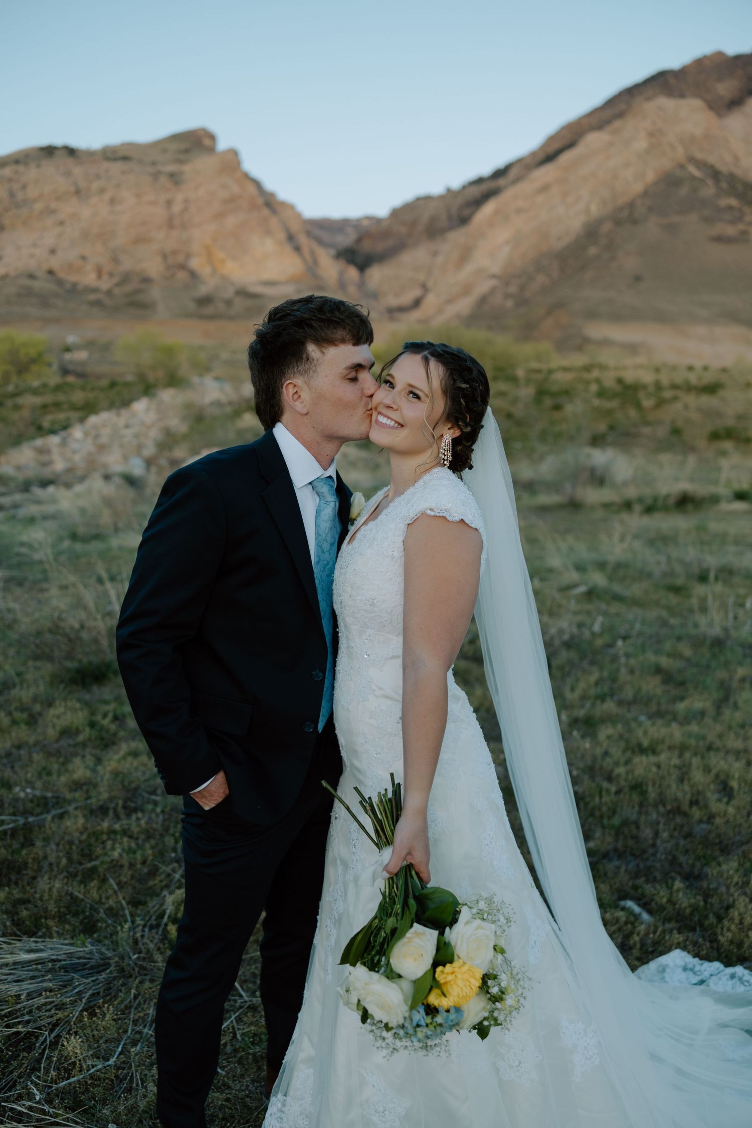 Groom in black suit kisses bride in white lace gown holding yellow and white bouquet with rocky mountain backdrop.