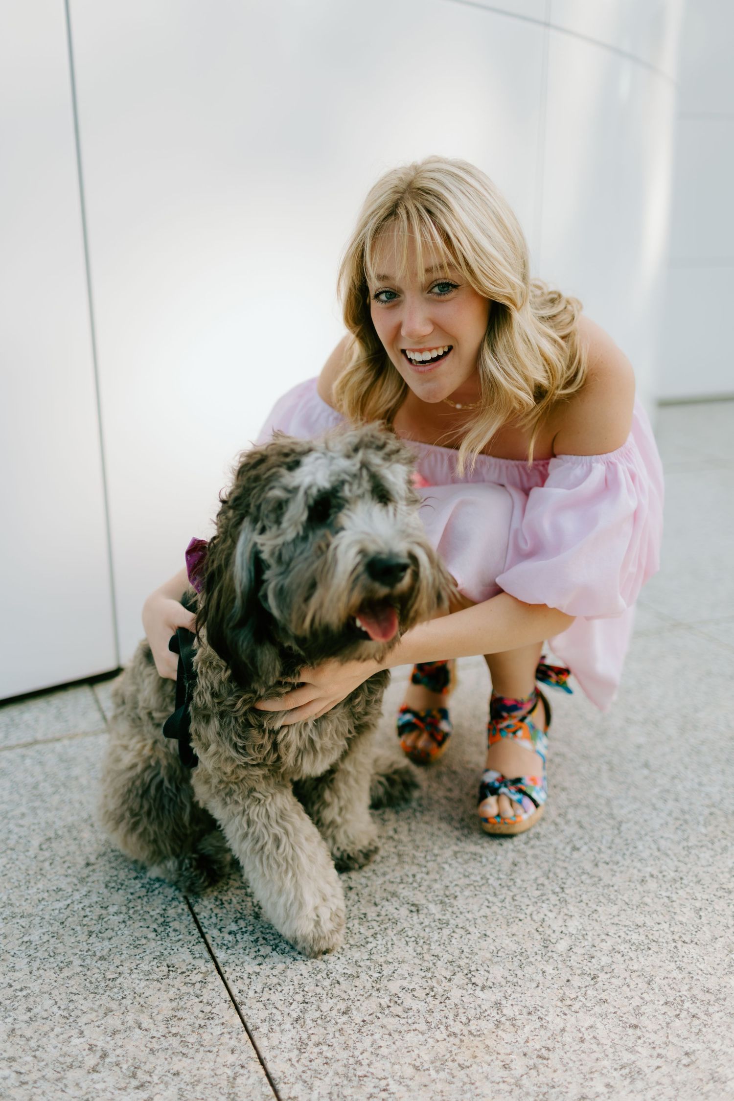 Smiling blonde woman in pink off-shoulder dress crouching with fluffy gray dog on tiled floor.