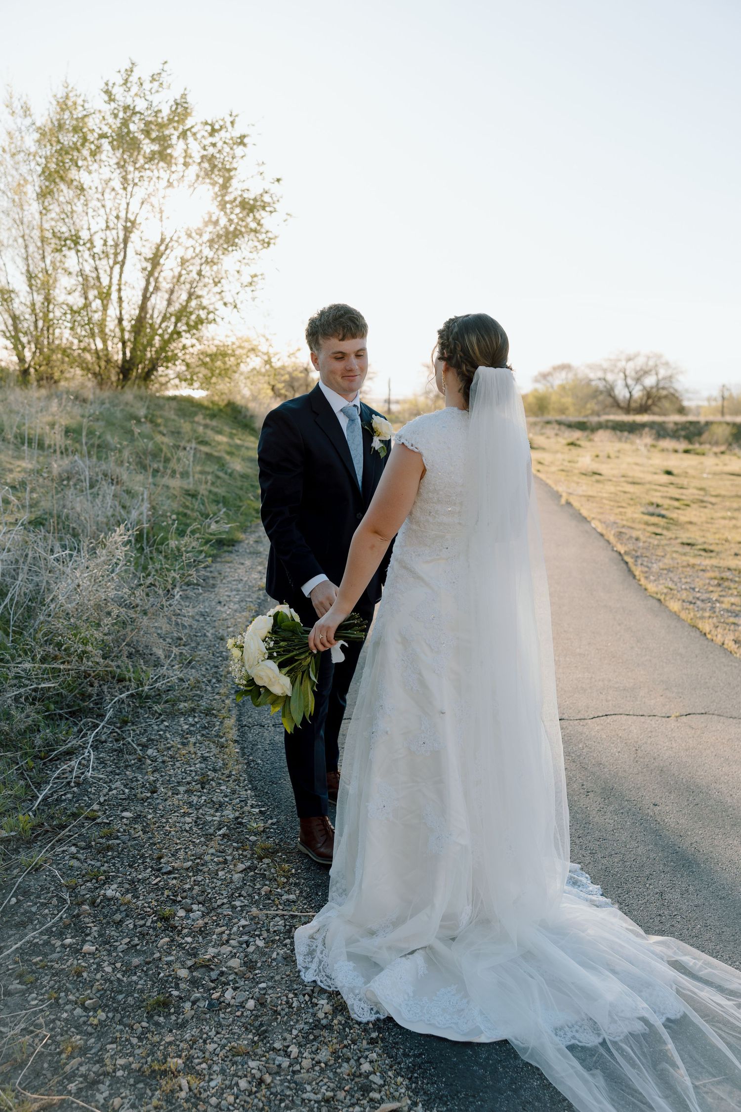 Bride in lace gown with veil and groom in black suit holding hands on sunlit path.