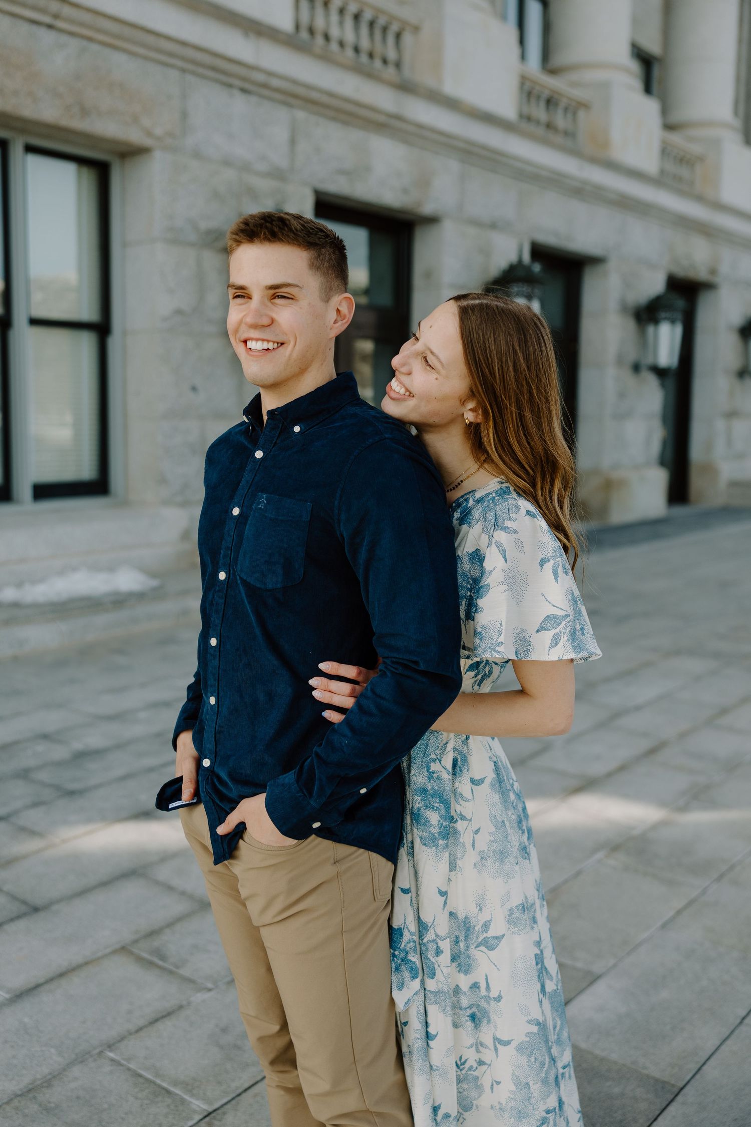 Smiling couple posing outdoors near a stone building, man in navy shirt and woman in floral dress.