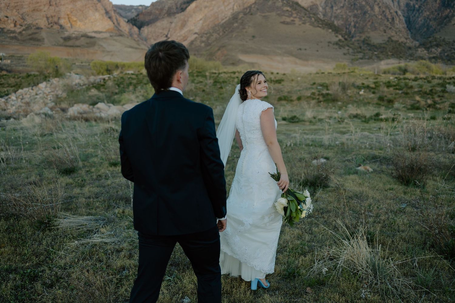 Bride and groom walk hand in hand through a rustic mountain meadow, bride holding a white floral bouquet.