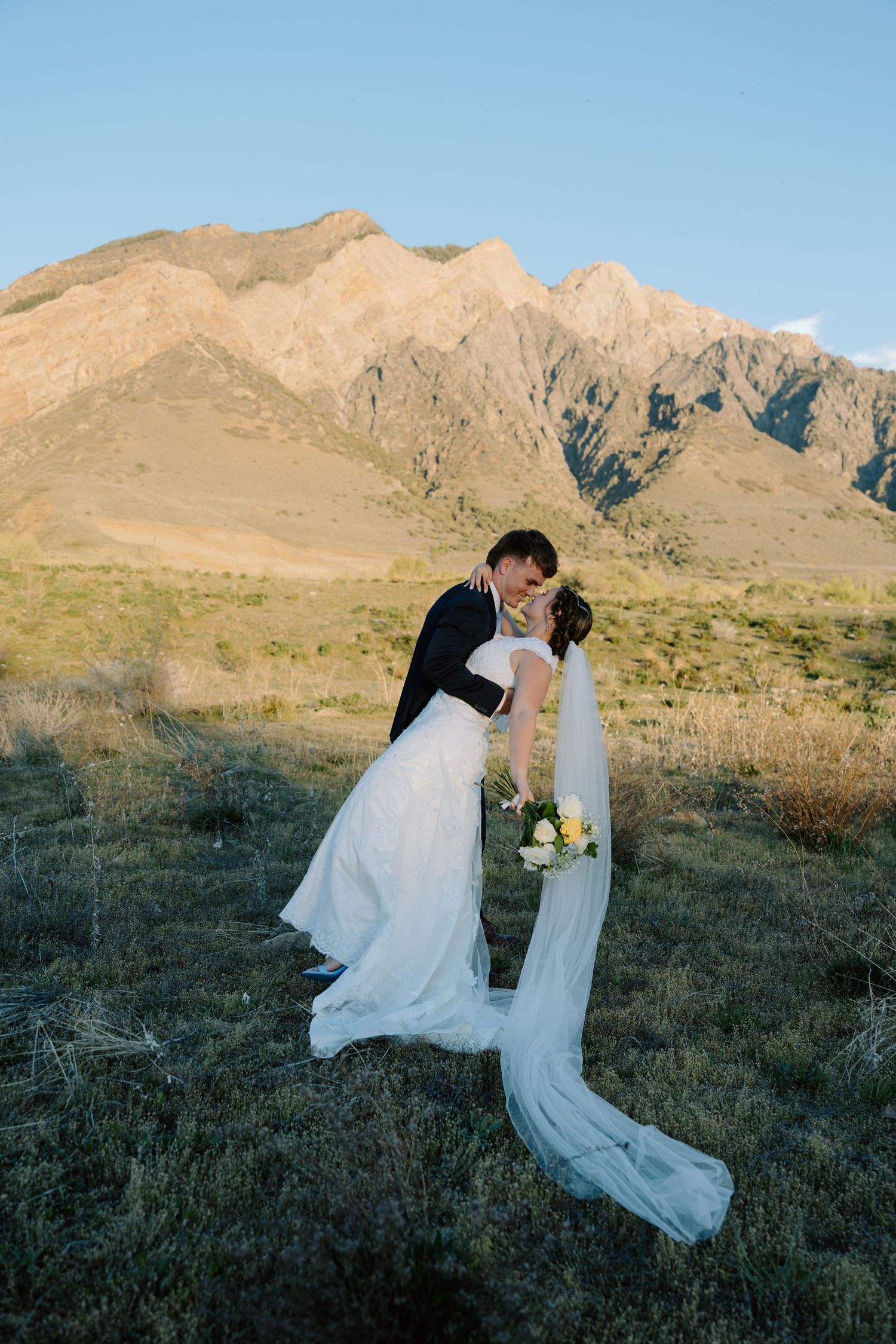 Groom dipping bride for a romantic kiss in a field with golden mountain backdrop.