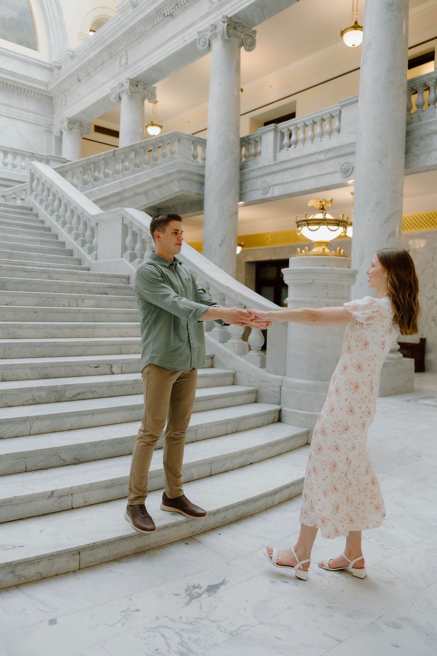 Couple holding hands and dancing playfully in elegant marble hall with grand staircase.