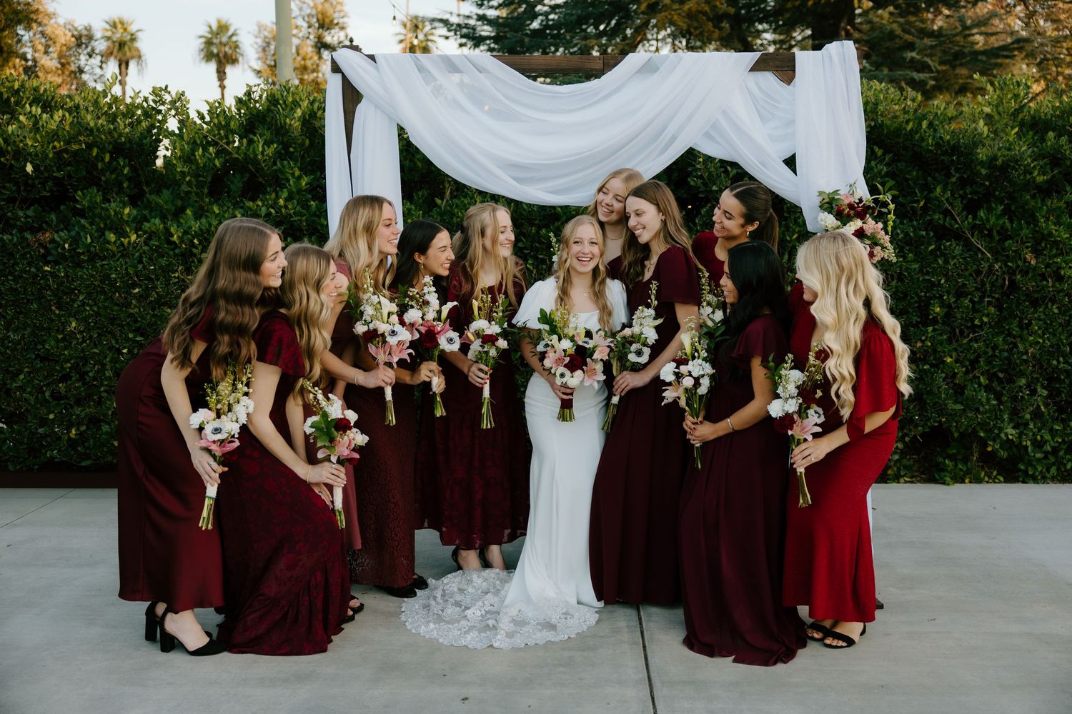 Bride in white lace gown surrounded by nine bridesmaids in burgundy dresses holding floral bouquets under white draped arch outdoors.