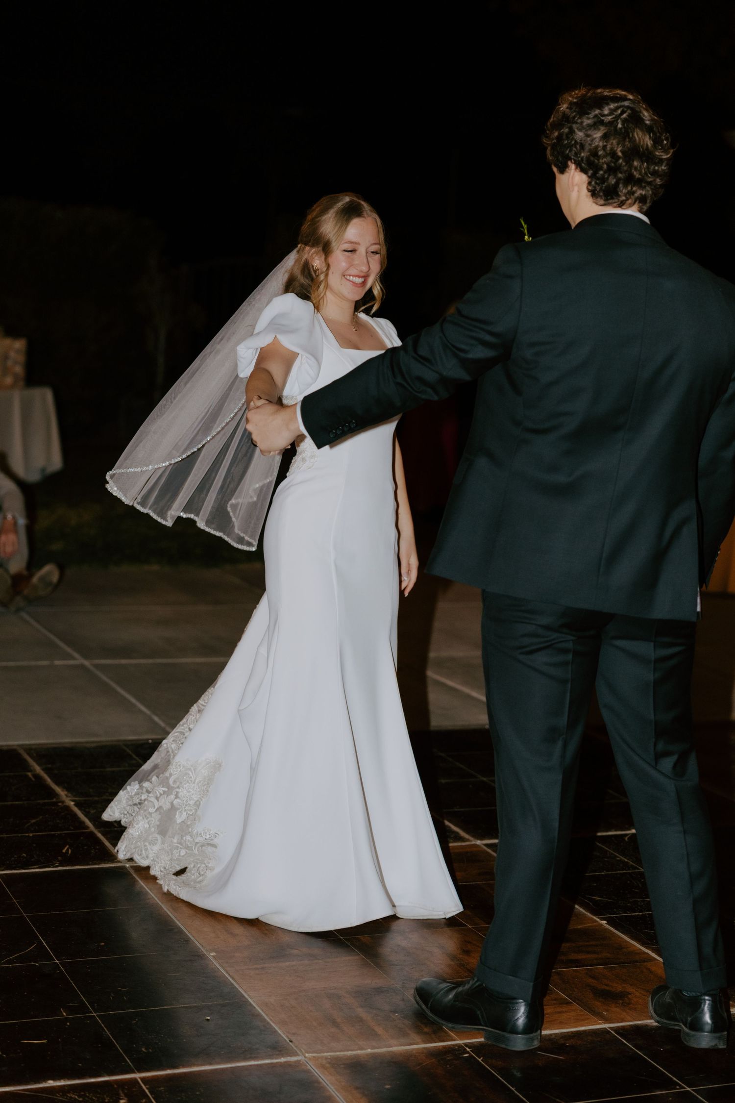 Bride in white mermaid gown with veil twirling during first dance with groom at night.