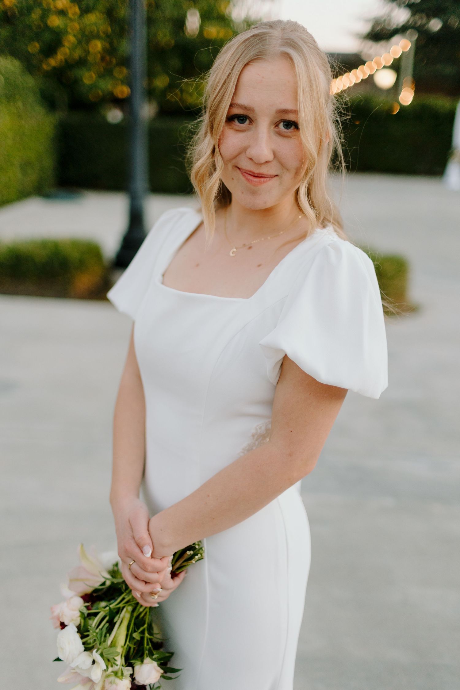 Elegant bride in white puff-sleeve wedding gown holds pink bouquet, smiling softly outdoors at golden hour.