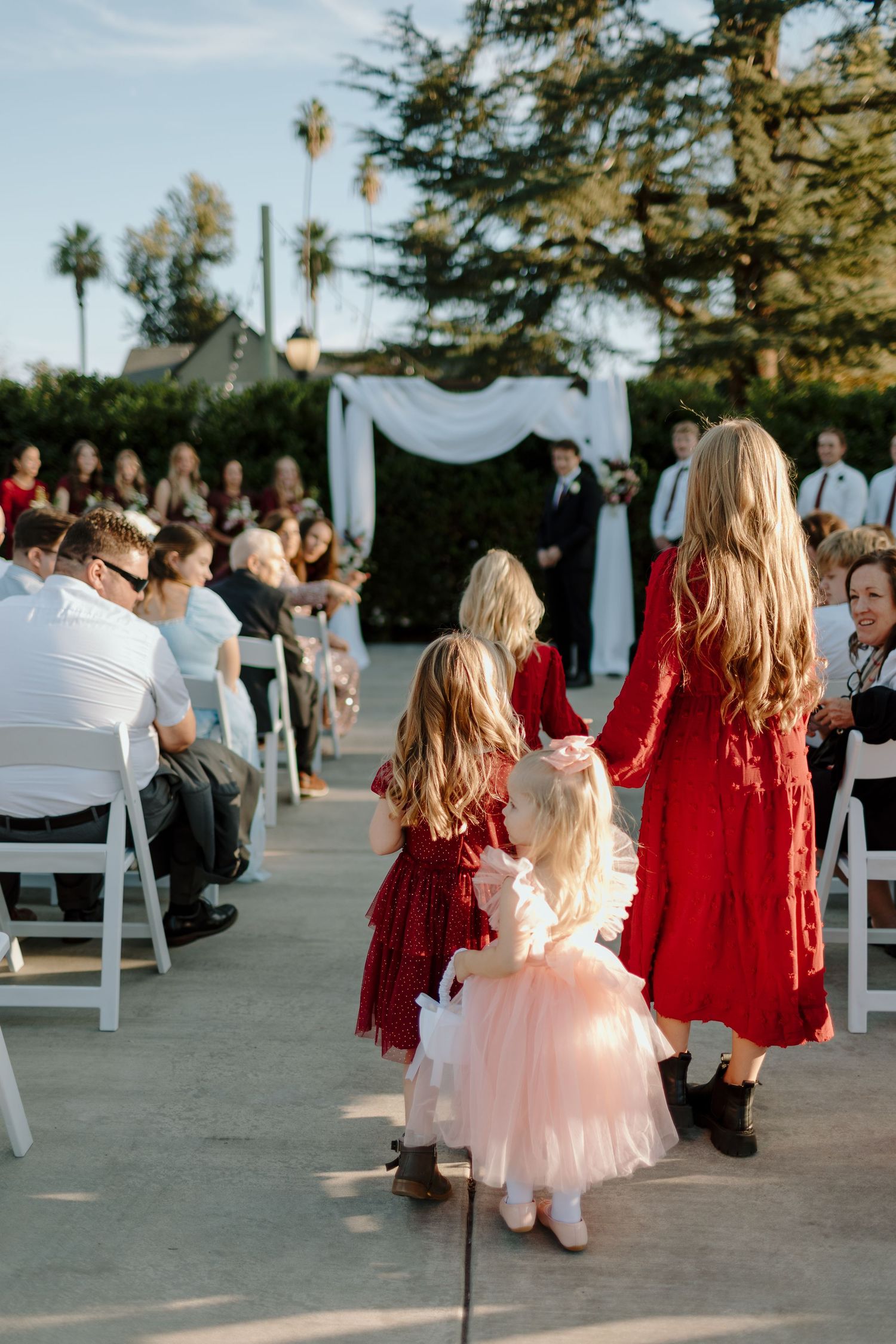 Flower girls in red and pink dresses walk down the outdoor wedding aisle toward a white floral arch ceremony backdrop.