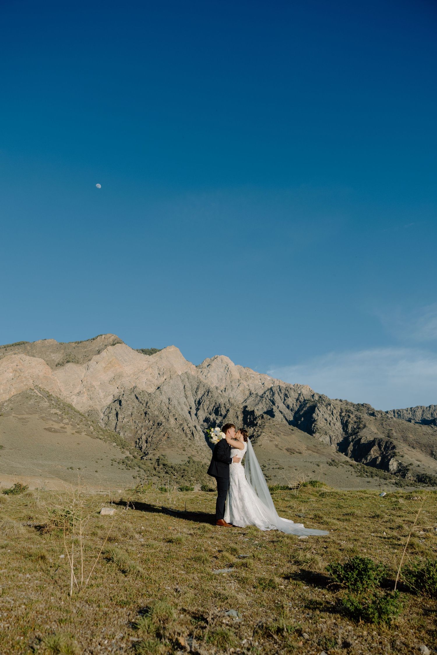 Bride and groom embracing on a mountain hilltop under a clear blue sky with rocky peaks behind them.