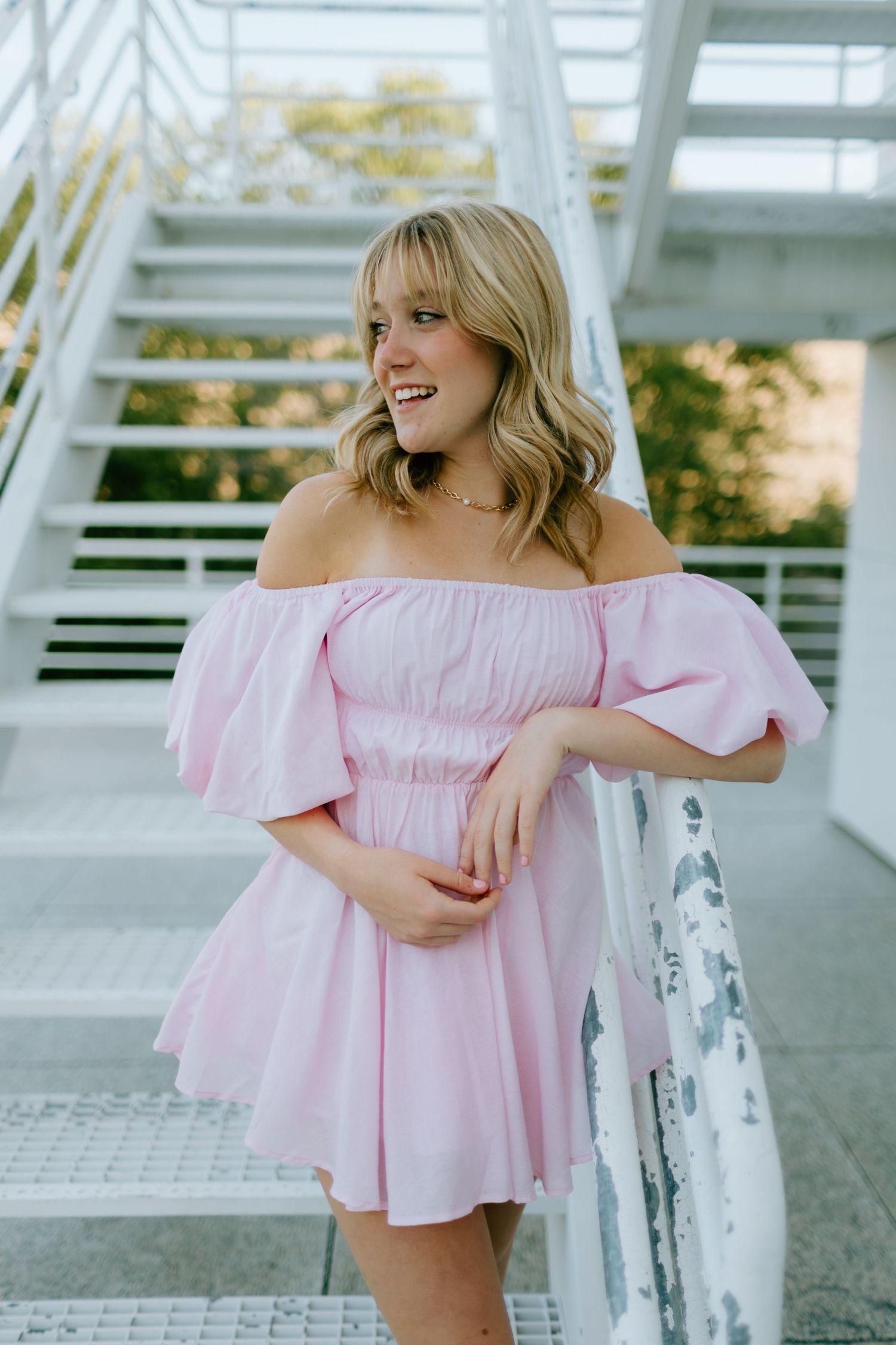 Young woman in pink off-shoulder puff sleeve mini dress posing on white metal stairs outdoors.