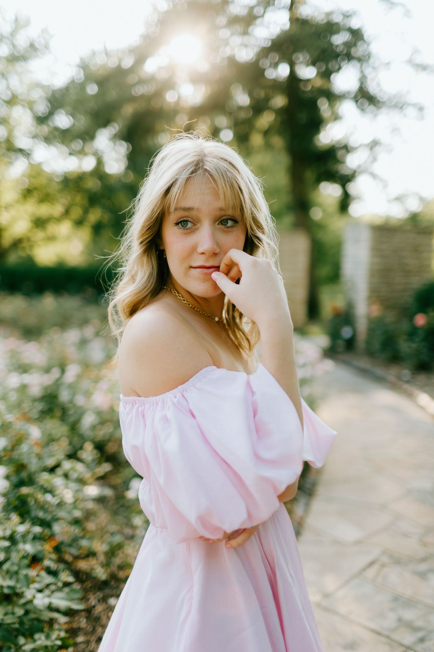 Blonde woman in pink off-shoulder dress posing in sunlit garden path.