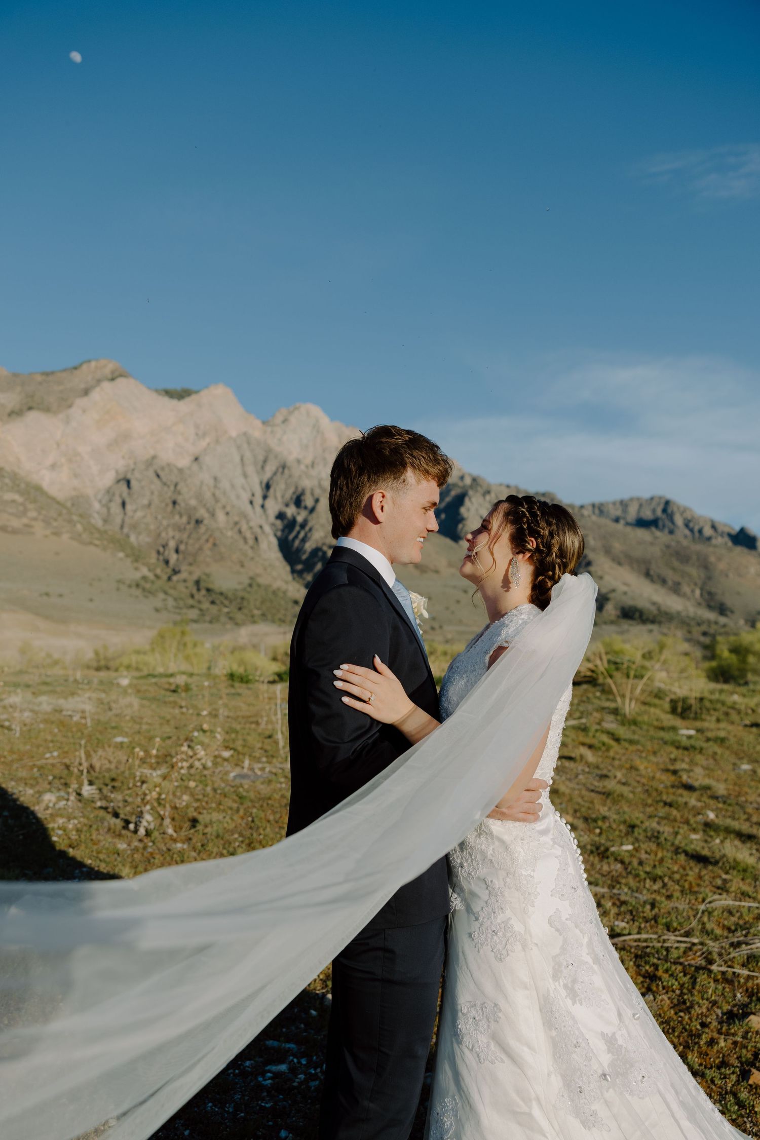 Groom embracing bride as her veil flows in wind with stunning mountain backdrop.