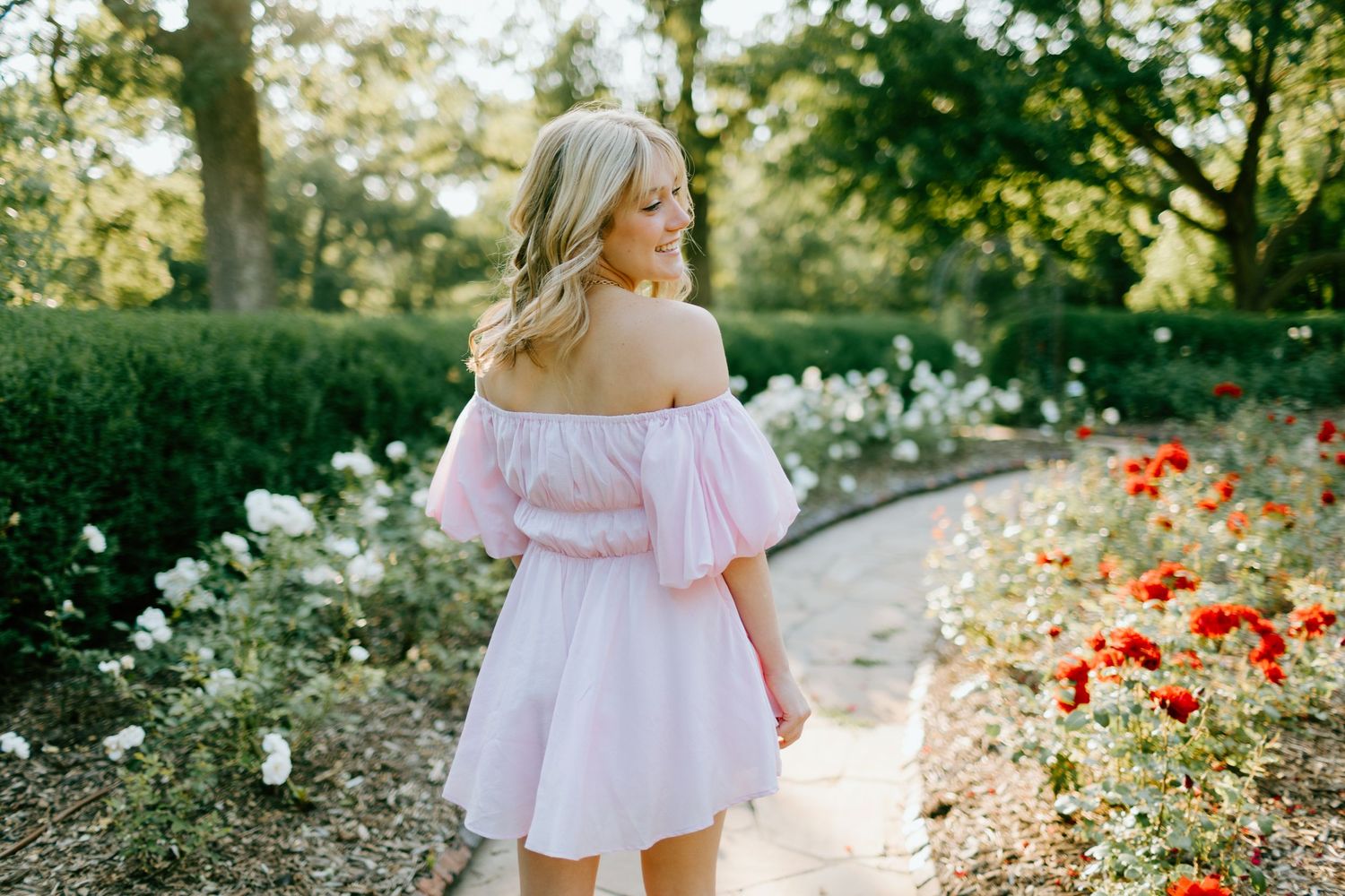 Blonde woman in pink off-shoulder dress walking garden path surrounded by red and white roses.