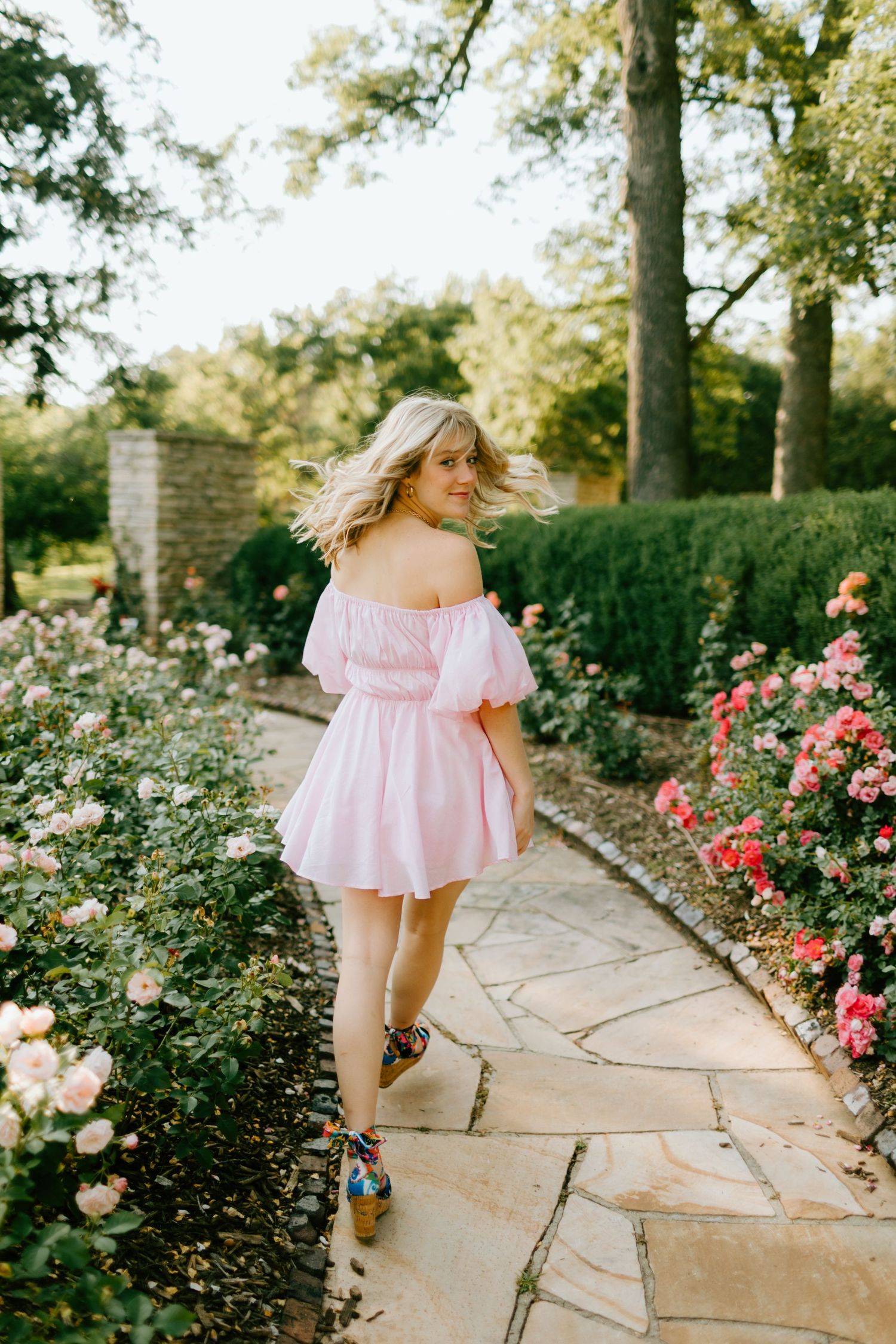 Blonde woman in pink off-shoulder mini dress walking stone garden path lined with blooming roses.