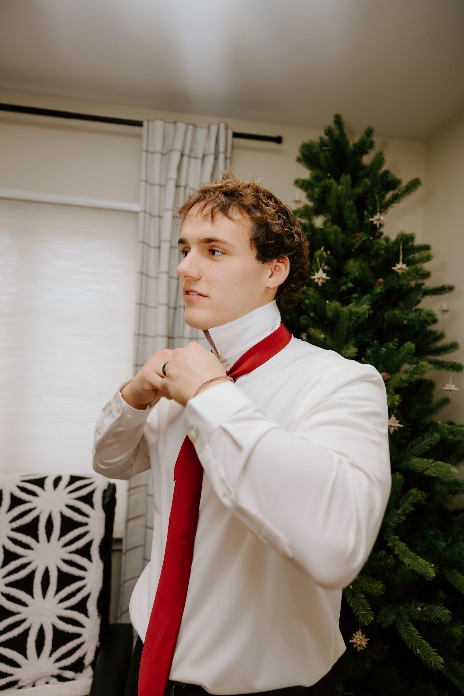 Groom in white shirt adjusting red tie near decorated Christmas tree before wedding ceremony.