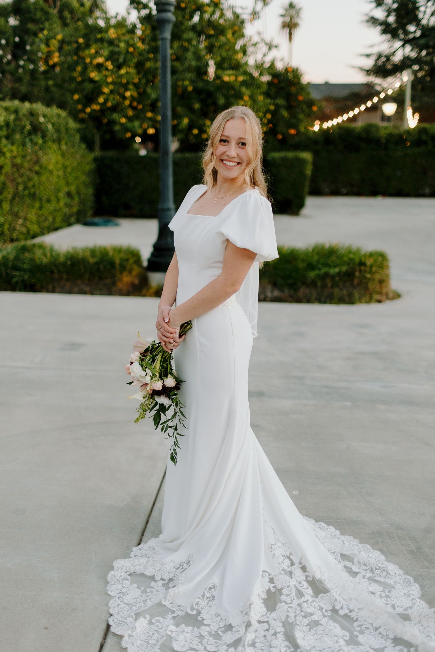 Smiling bride in elegant white flutter-sleeve gown with lace train holds a pink and white bouquet outdoors.