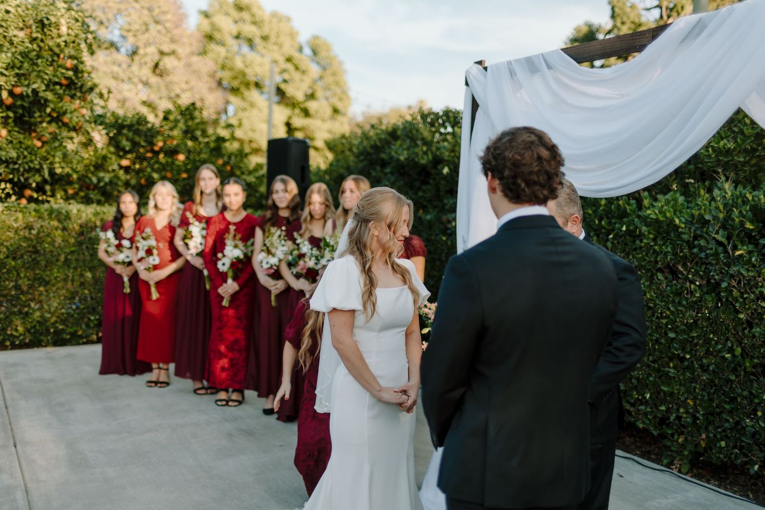 Bride and groom stand at outdoor wedding altar with bridesmaids in red dresses holding bouquets under draped canopy.