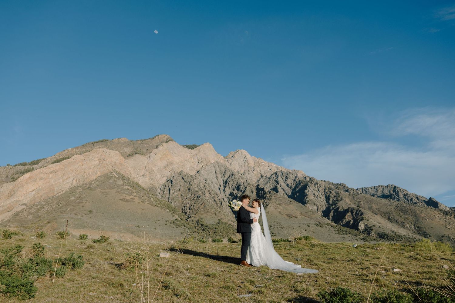 Wedding couple embracing on a scenic mountain landscape under a clear blue sky with rugged peaks behind them.