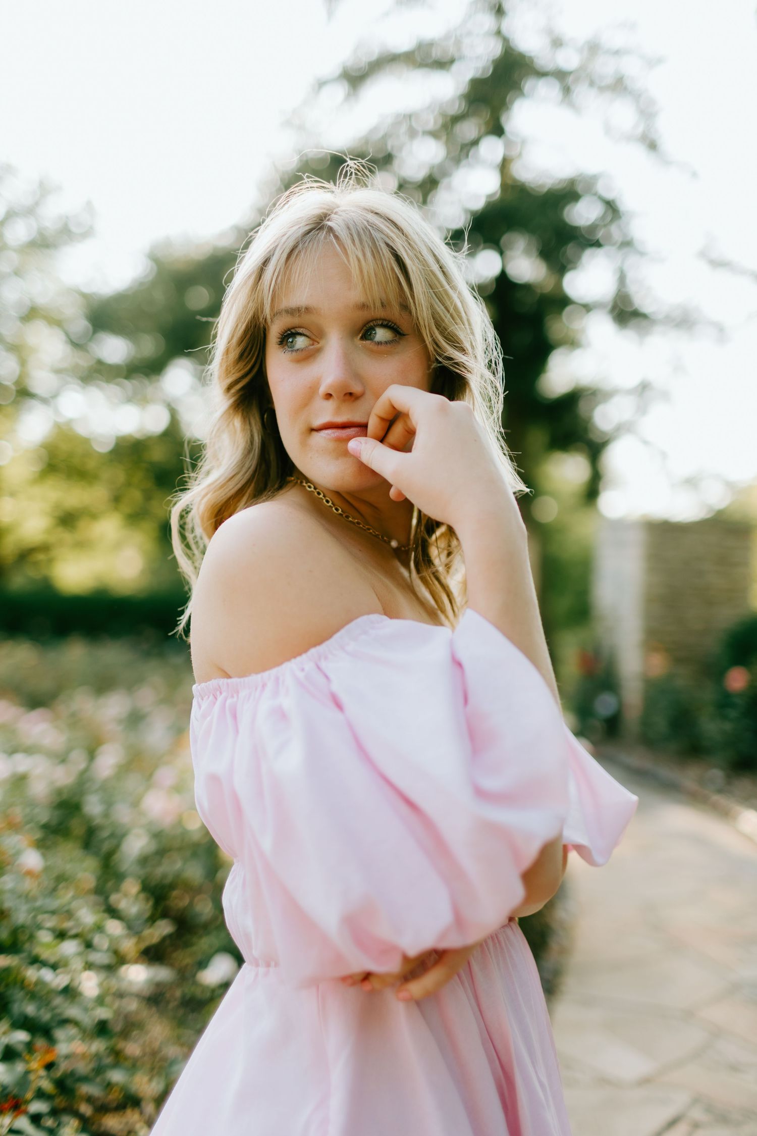 Young blonde woman in pink off-shoulder dress posing thoughtfully in a sunlit garden.
