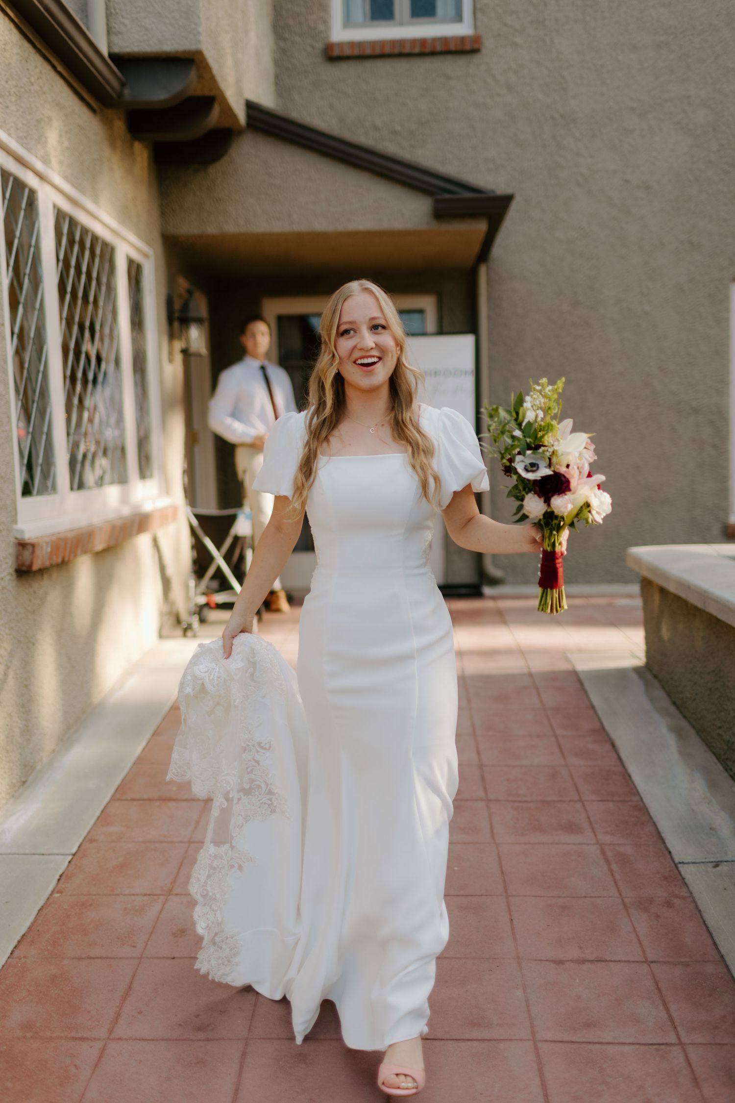 Smiling bride in white square-neck gown holding floral bouquet walks along outdoor path on wedding day.