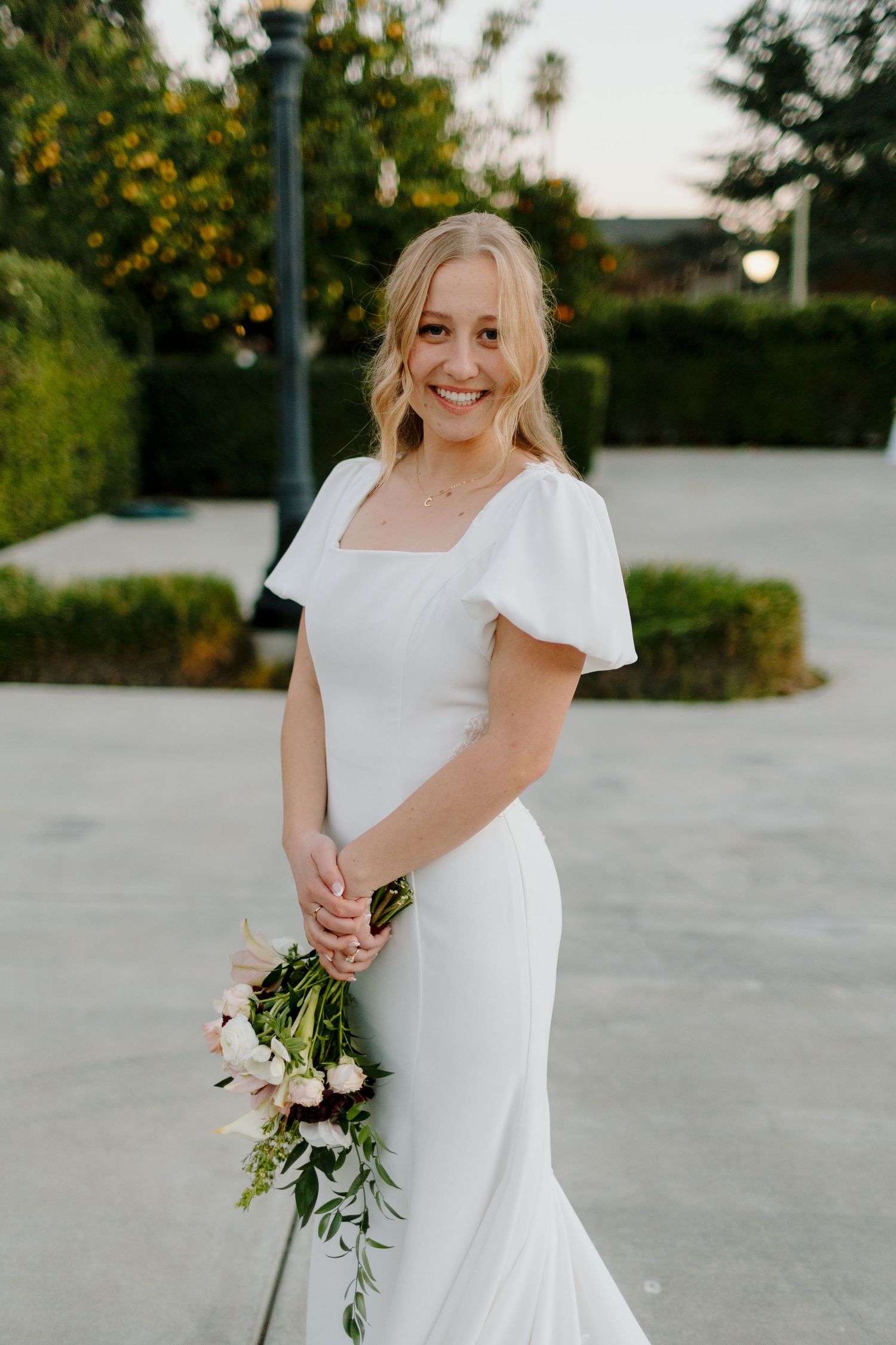 Smiling bride in white flutter-sleeve wedding gown holds a bouquet of blush flowers and greenery outdoors.