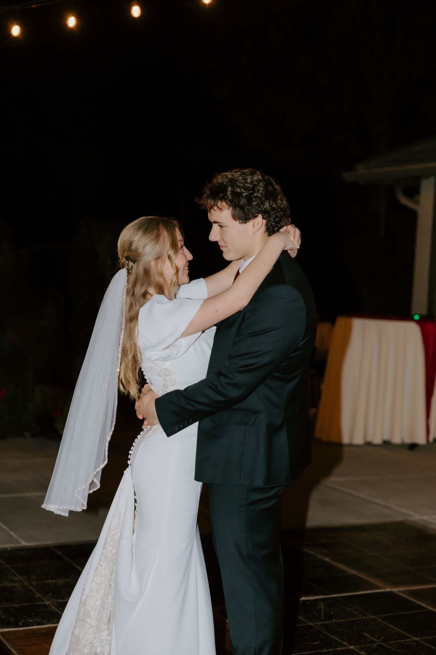Bride and groom sharing intimate first dance outdoors at night under string lights.