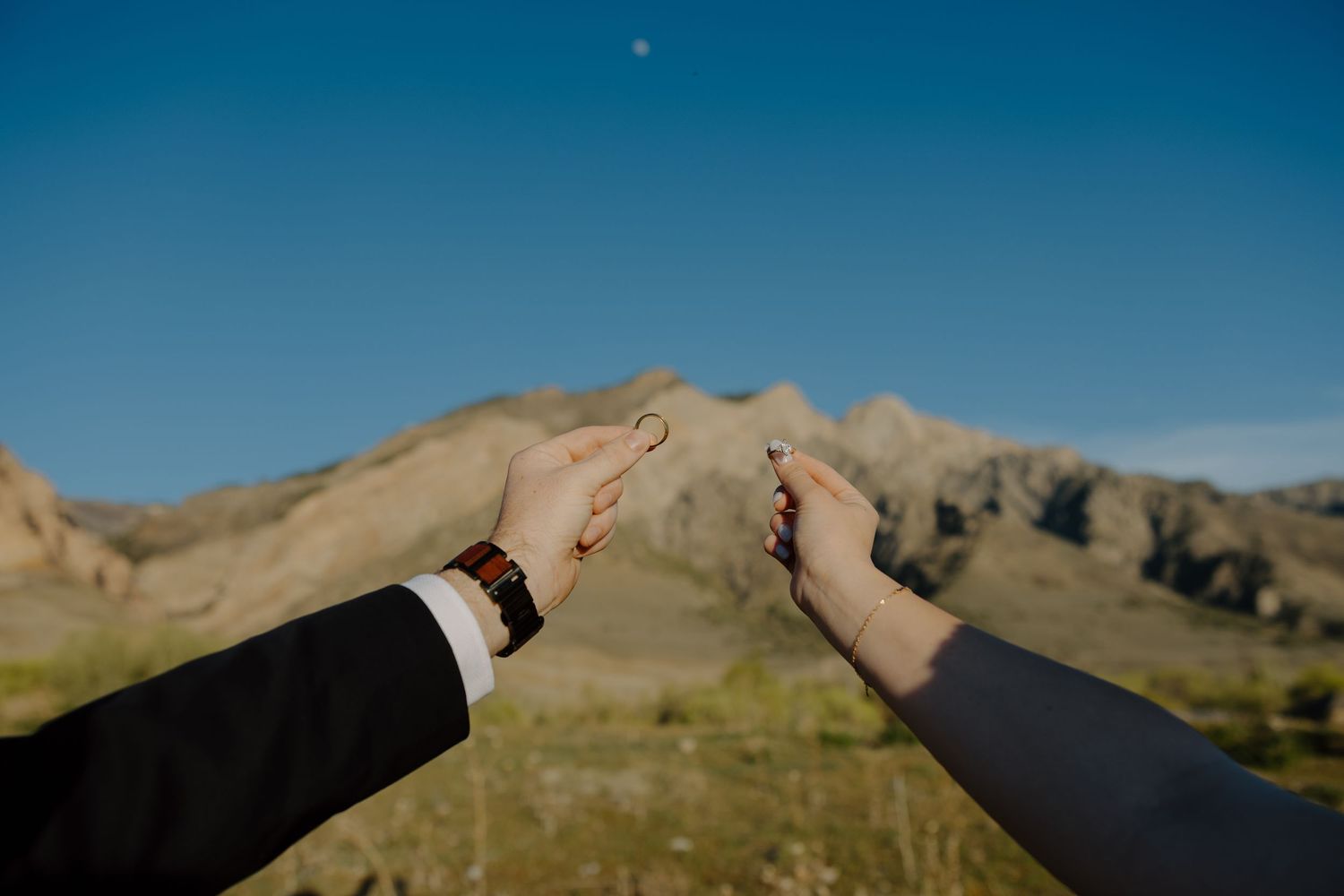 Bride and groom hold up wedding rings against a scenic mountain and blue sky backdrop.