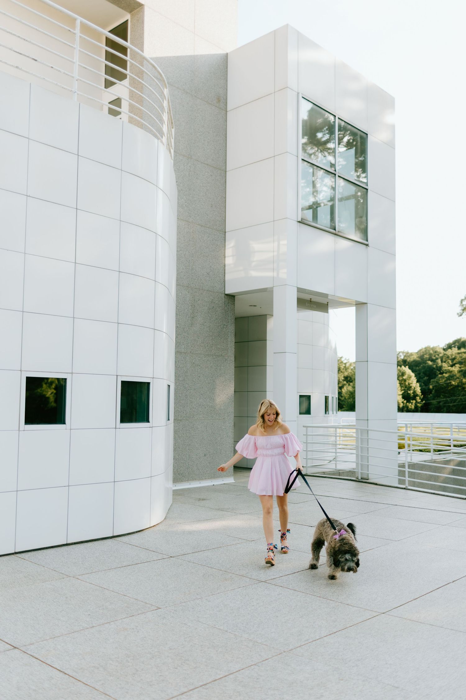 Woman in pink off-shoulder dress walking small dog on leash outside modern white building on sunny day.
