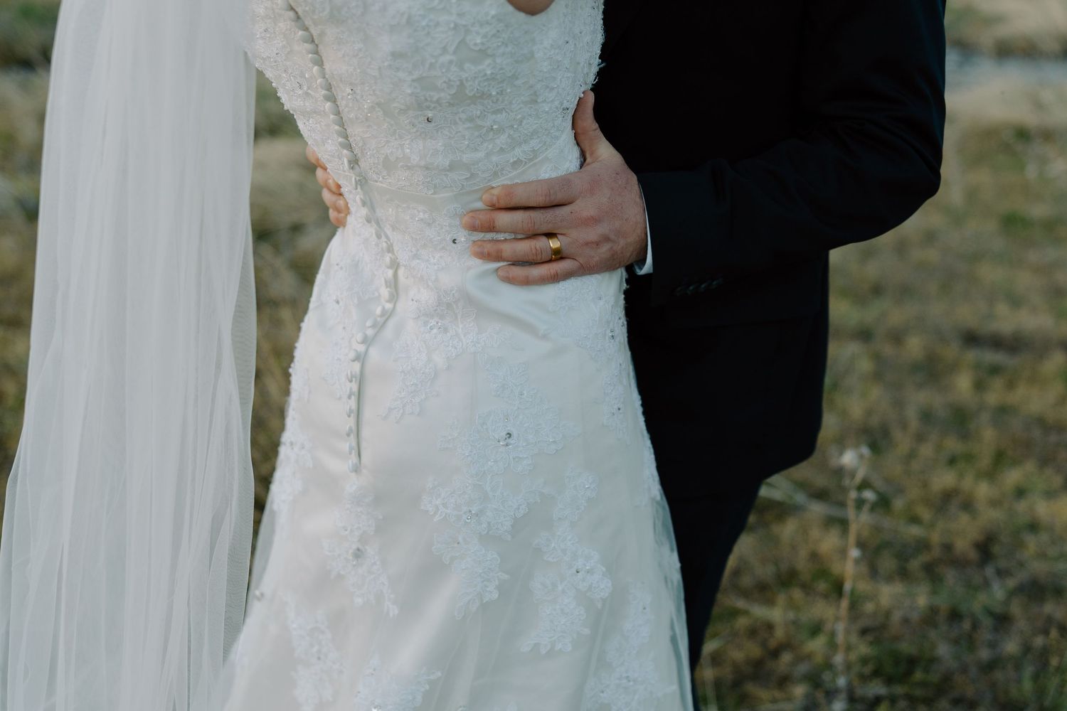 Close-up of bride in lace wedding gown with groom in black suit embracing outdoors.