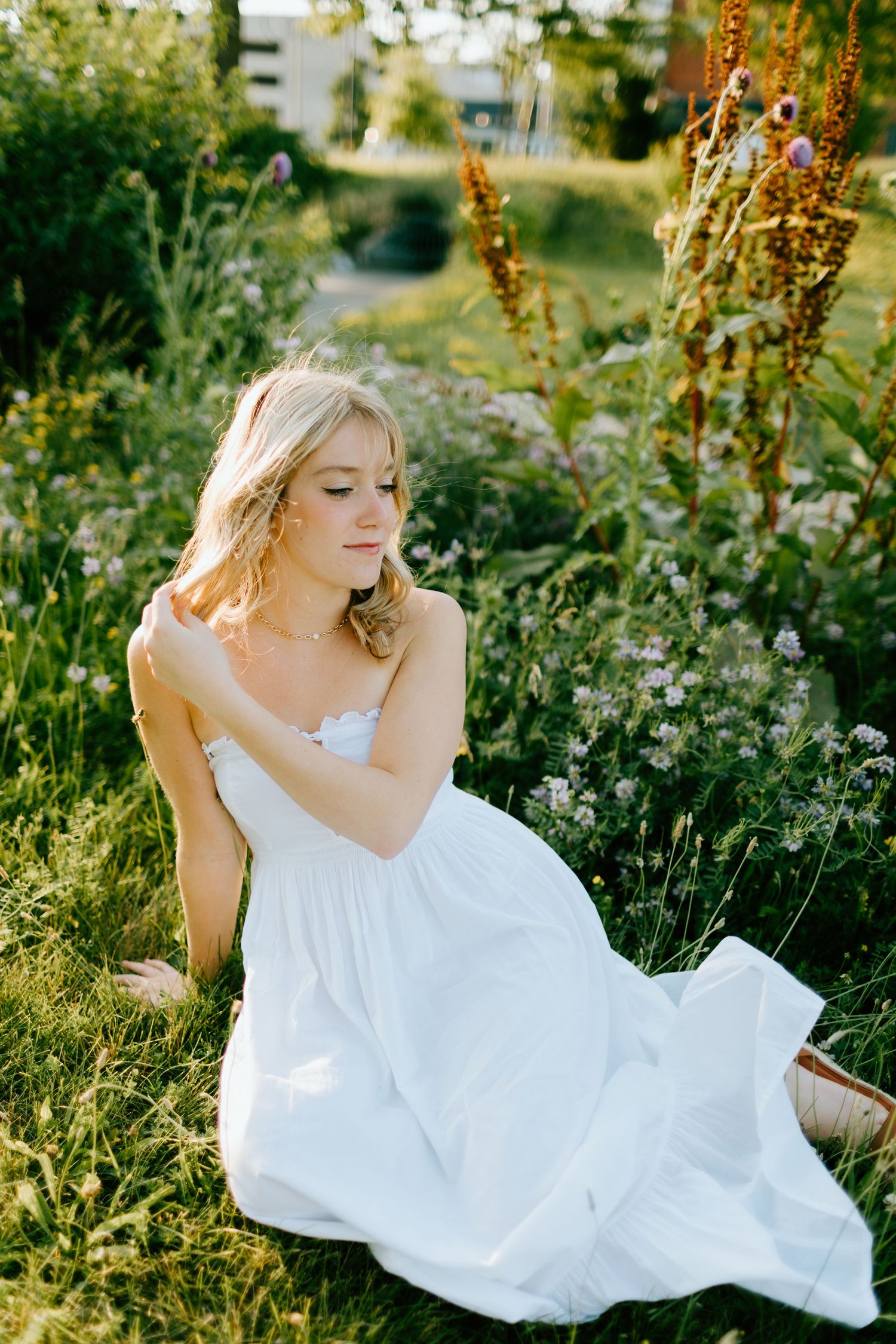 Blonde woman in flowing white dress sitting in lush green meadow with wildflowers in warm sunlight.