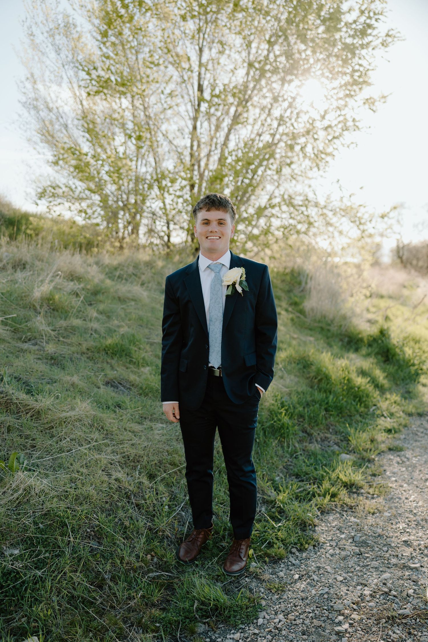 Young groom in dark suit with white boutonniere smiling outdoors on a sunny day.