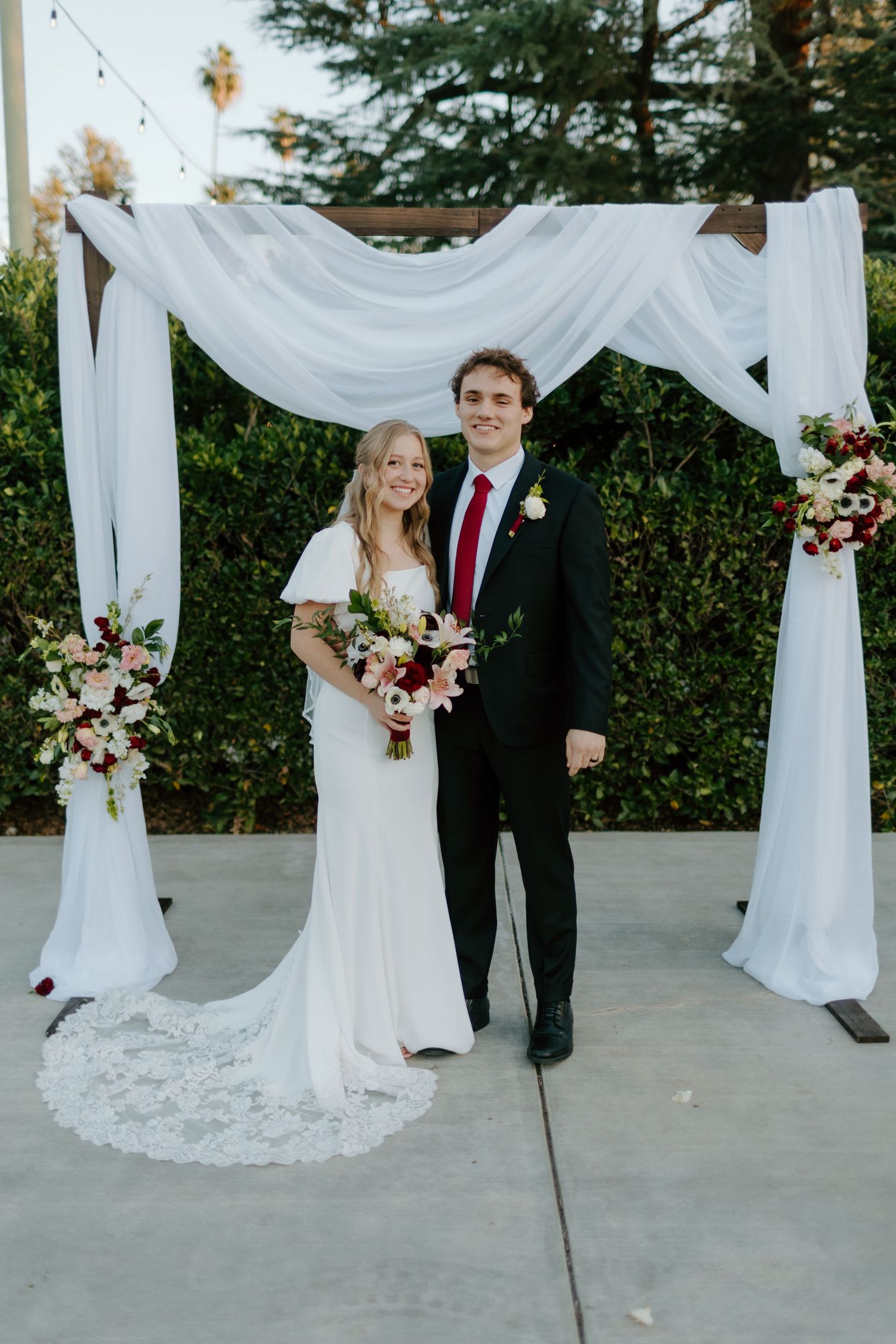 Bride and groom smiling under white draped wedding arch with burgundy floral arrangements outdoors.