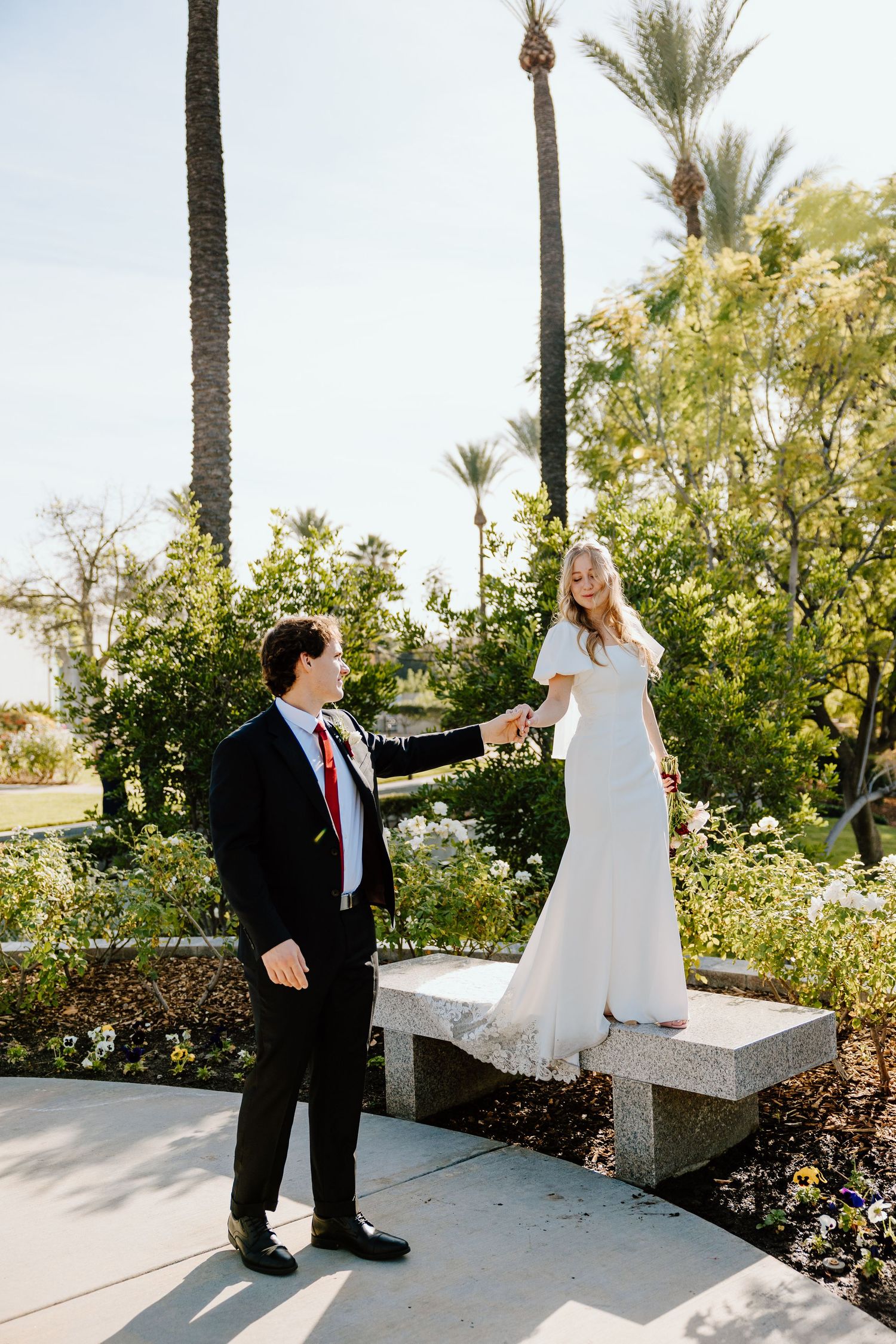 A bride in a white gown holds hands with a groom in a black suit amid lush greenery and palm trees outdoors.