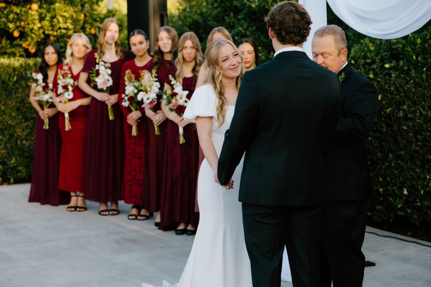 Bride and groom exchange vows at outdoor ceremony while bridesmaids in burgundy dresses stand alongside.