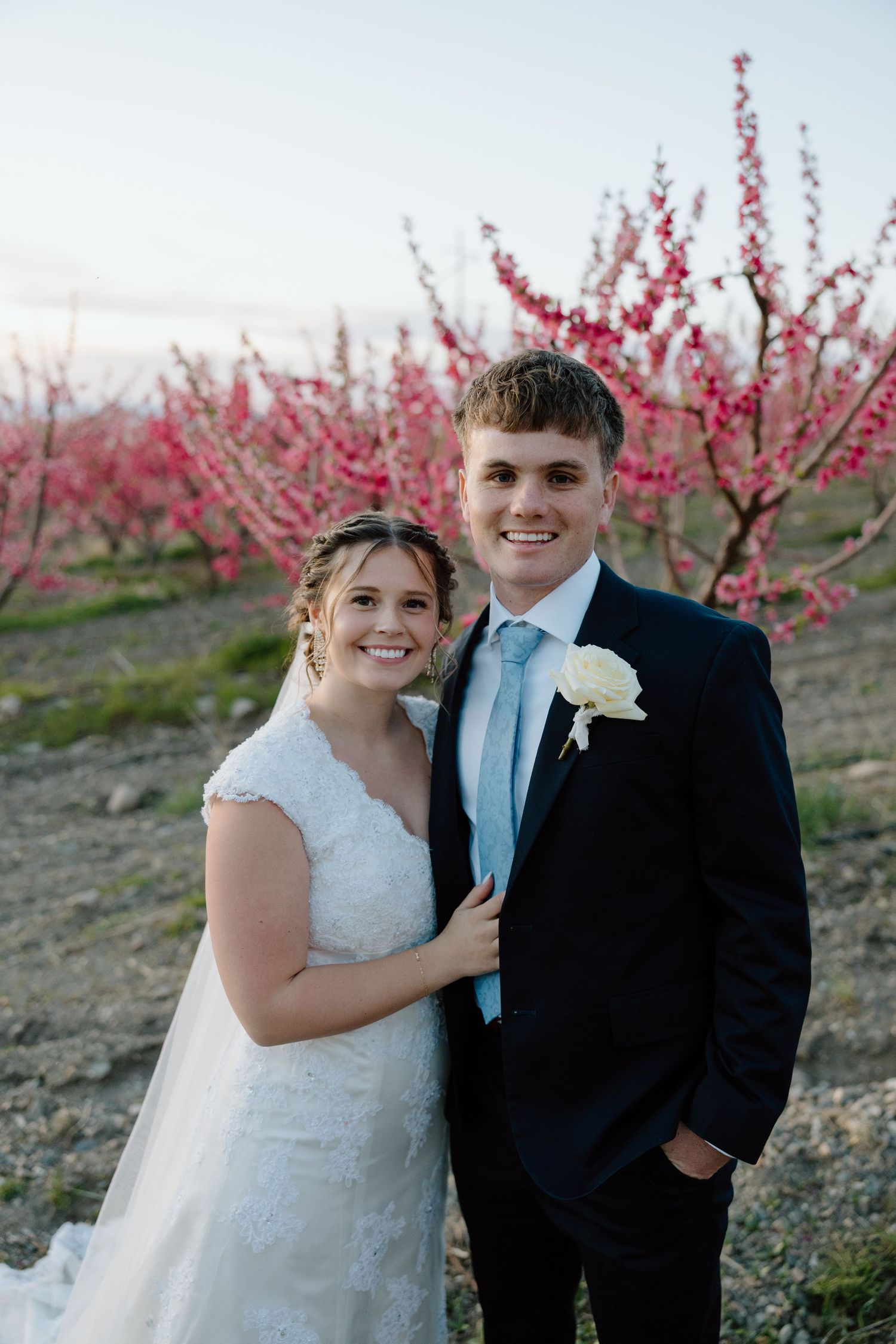Bride and groom smiling together in front of blooming pink cherry trees at an outdoor spring wedding.