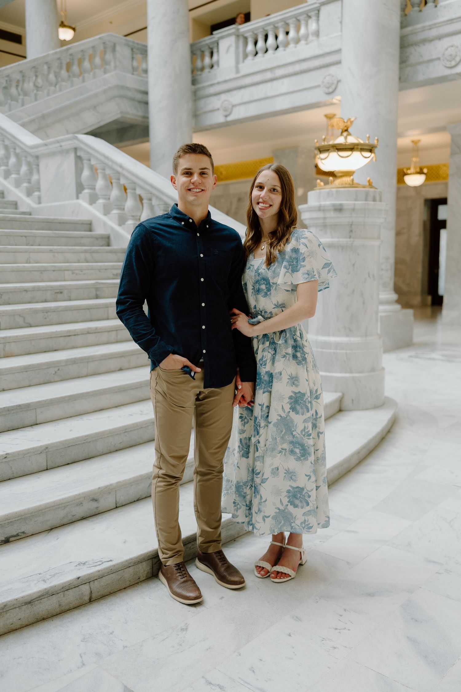 Young couple posing together inside elegant marble building with grand staircase and chandeliers.