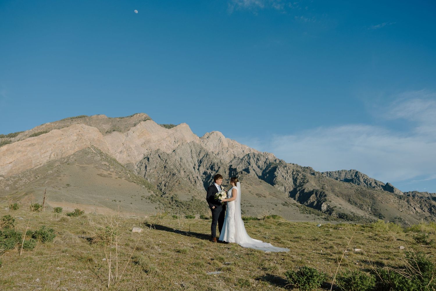 Bride and groom stand together on a mountain hillside under a clear blue sky during their outdoor wedding ceremony.