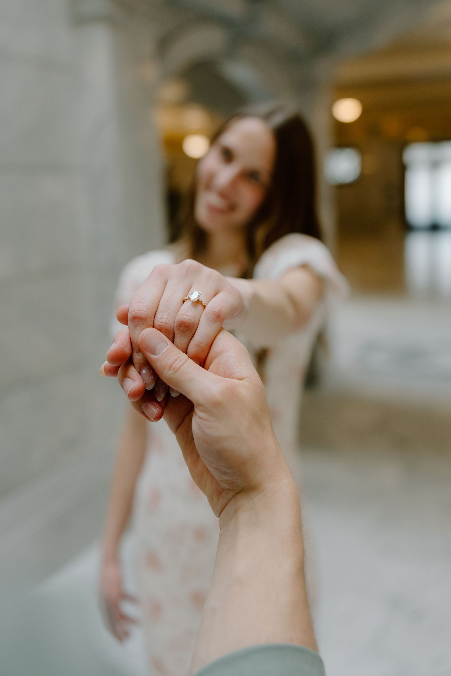 A close-up of a woman extending her hand displaying an engagement ring toward the camera in a marble hall.