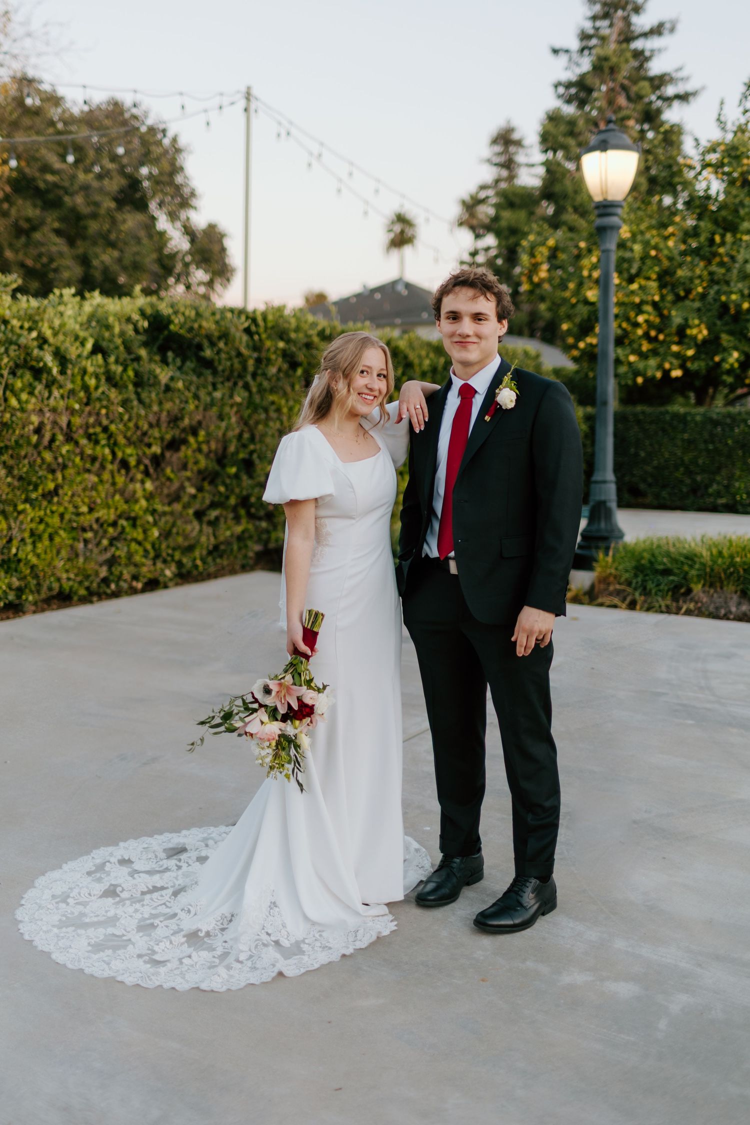 Bride in white flutter-sleeve gown with lace train stands with groom in black suit and red tie at outdoor venue.