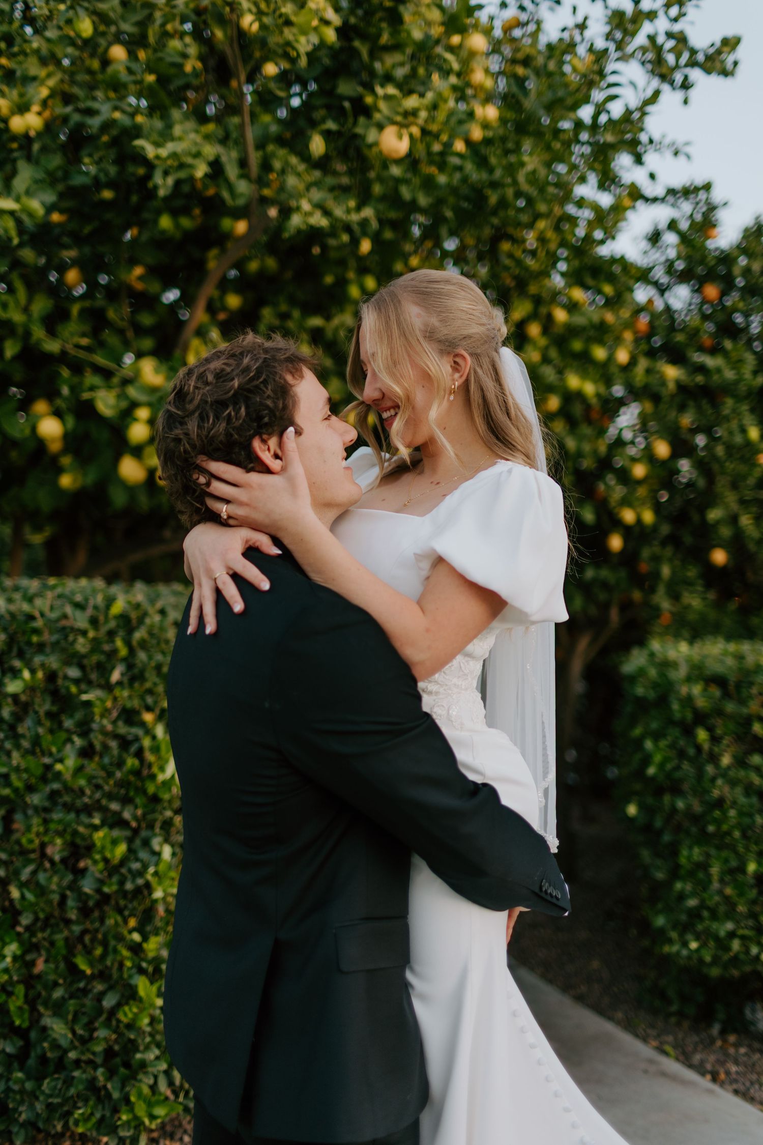 Bride and groom embracing and smiling near a lemon tree in an outdoor garden setting.