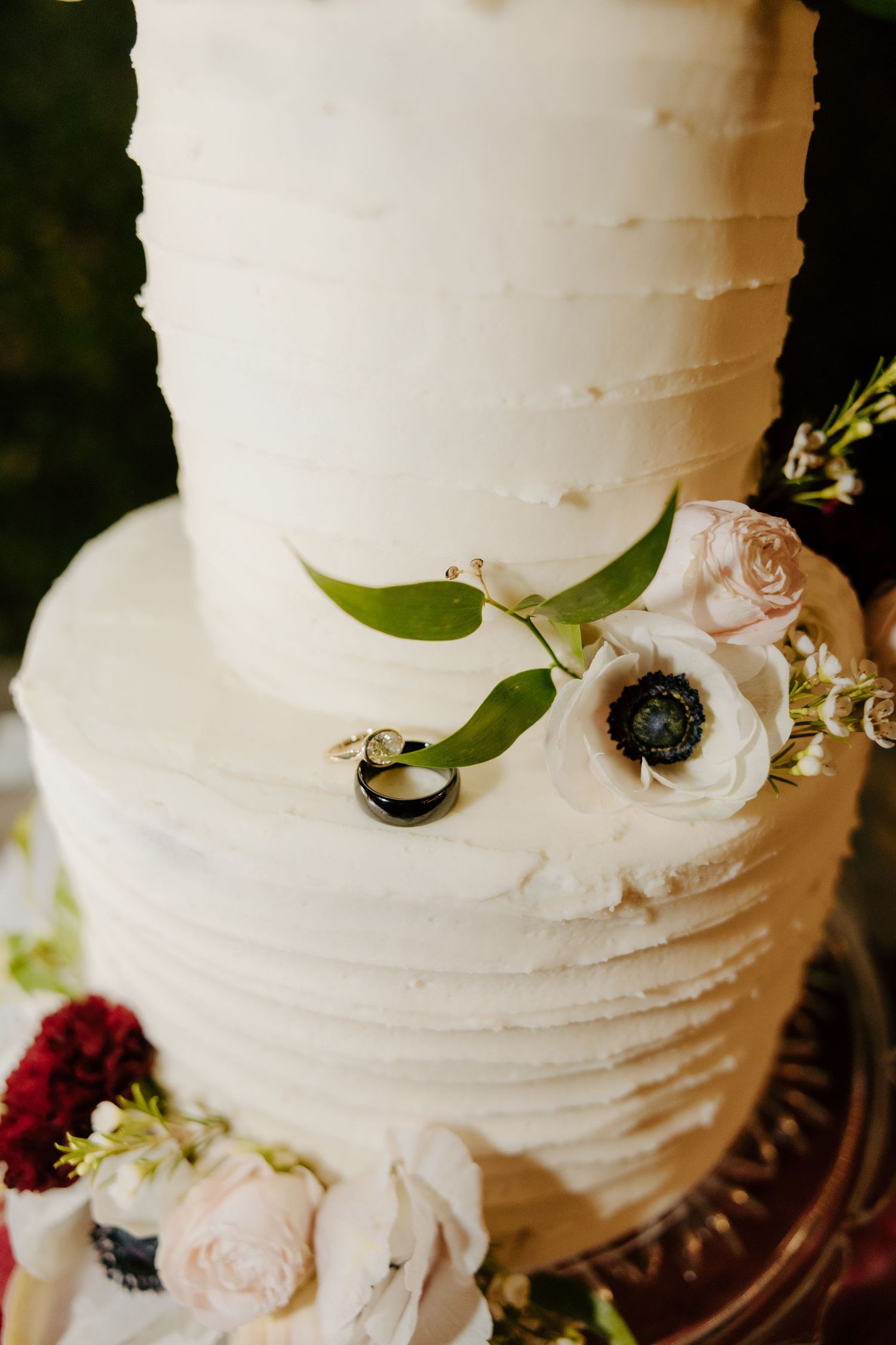 Two-tier white textured wedding cake decorated with anemone flowers, roses, greenery, and two wedding rings.