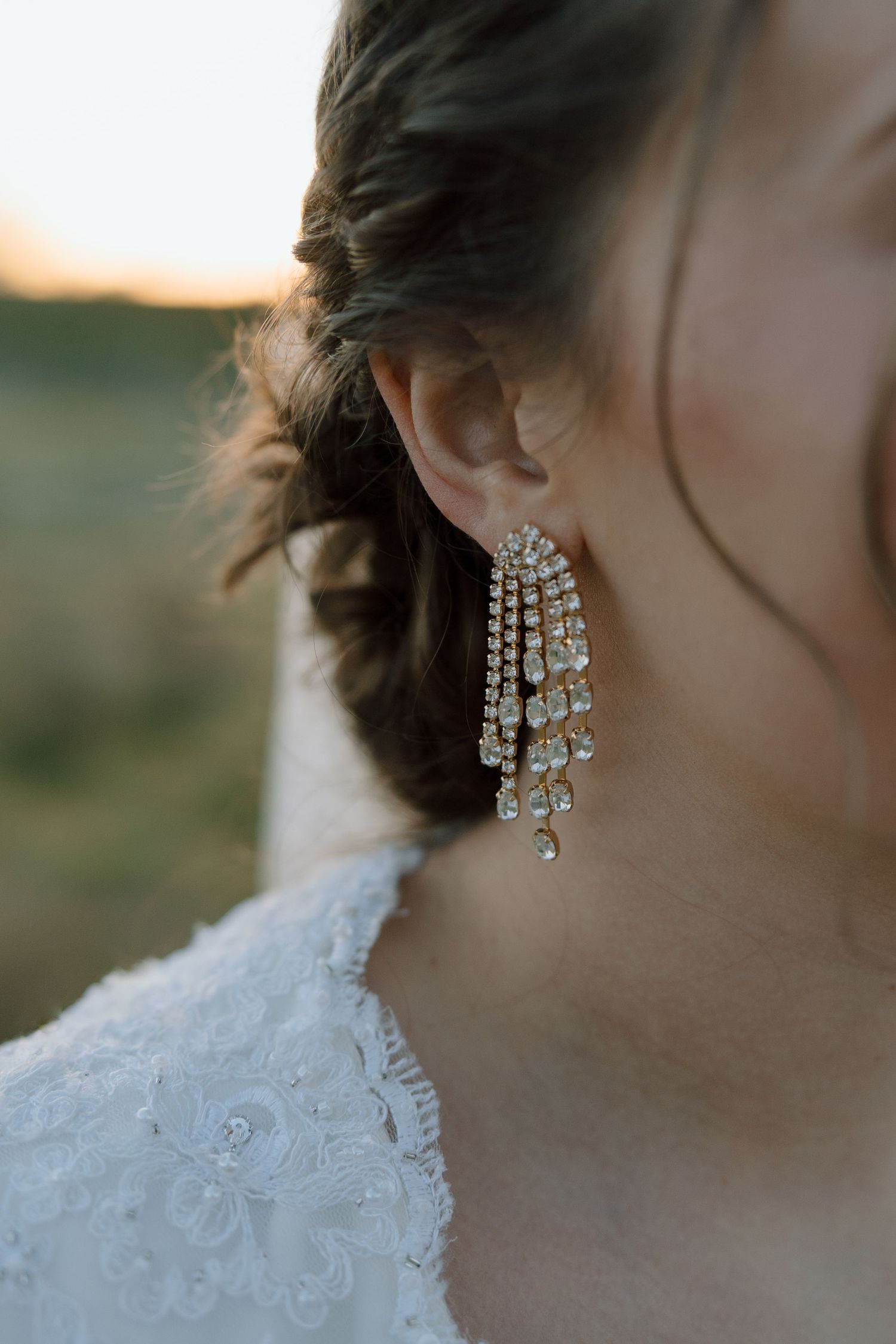 Close-up of a bride wearing elegant crystal chandelier drop earrings with lace wedding dress at golden hour.