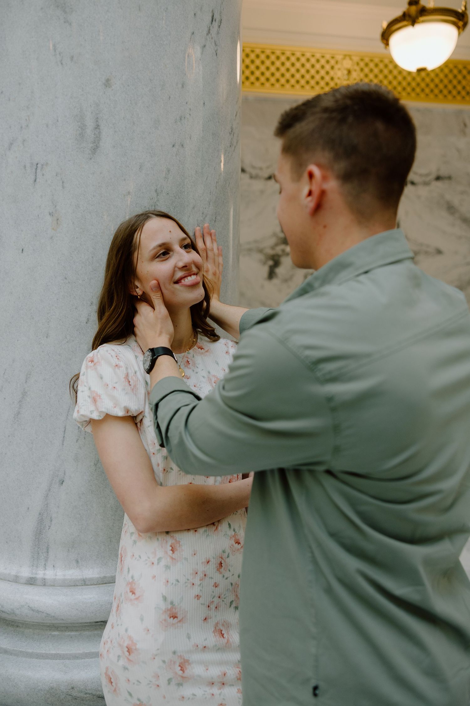 A man in a green shirt gently touches a smiling woman's face near marble columns in an elegant setting.