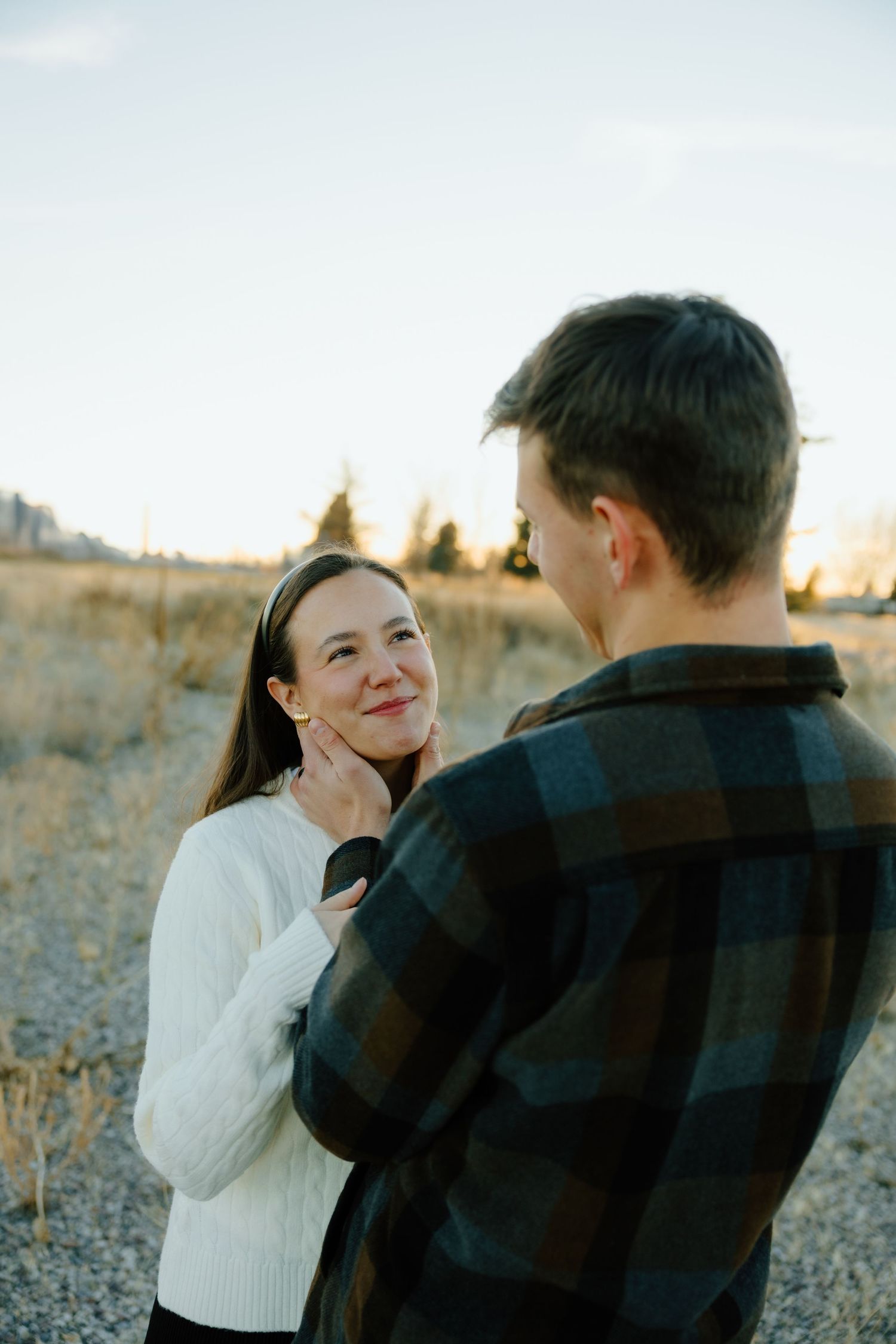 A couple shares a tender moment outdoors, woman in white sweater smiling at man in plaid shirt against golden field backdrop.