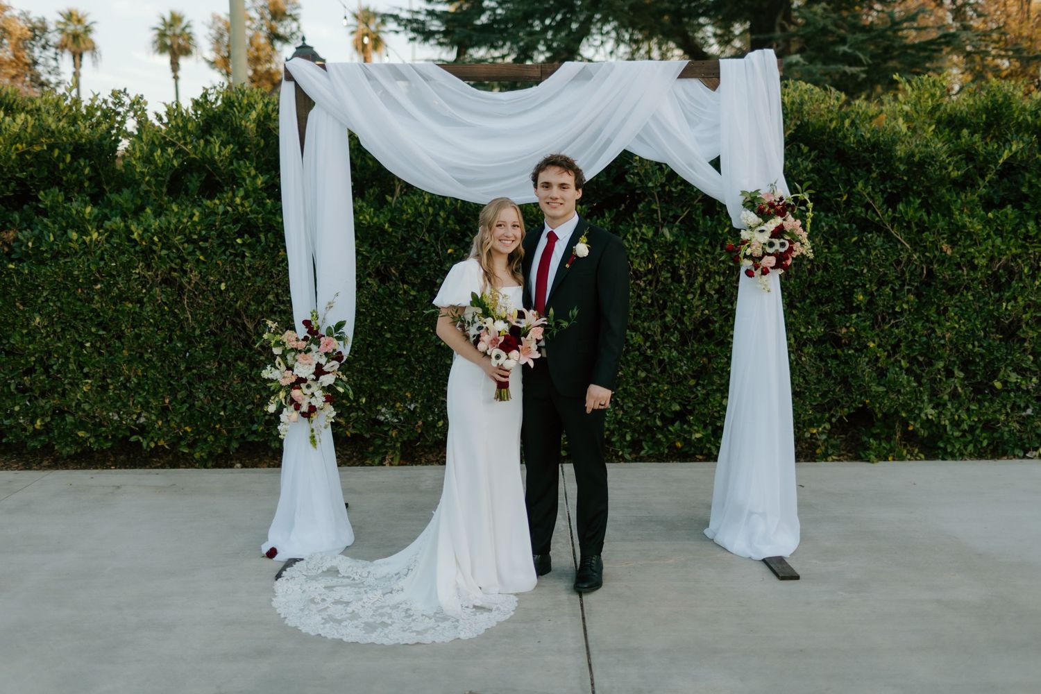 Bride and groom smile together under white draped arch adorned with red and pink floral arrangements outdoors.